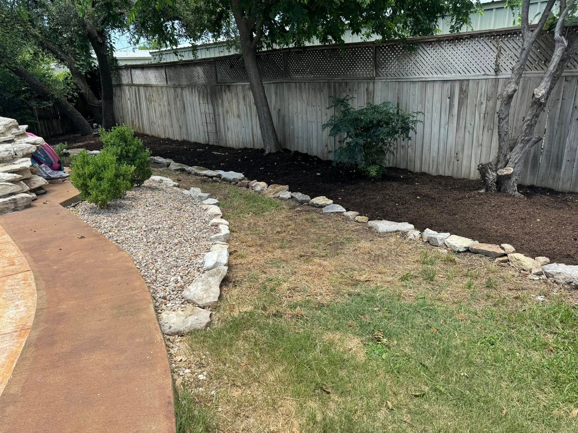 A backyard with a stone path, landscaping, and a wooden fence. Some brown mulch is present.