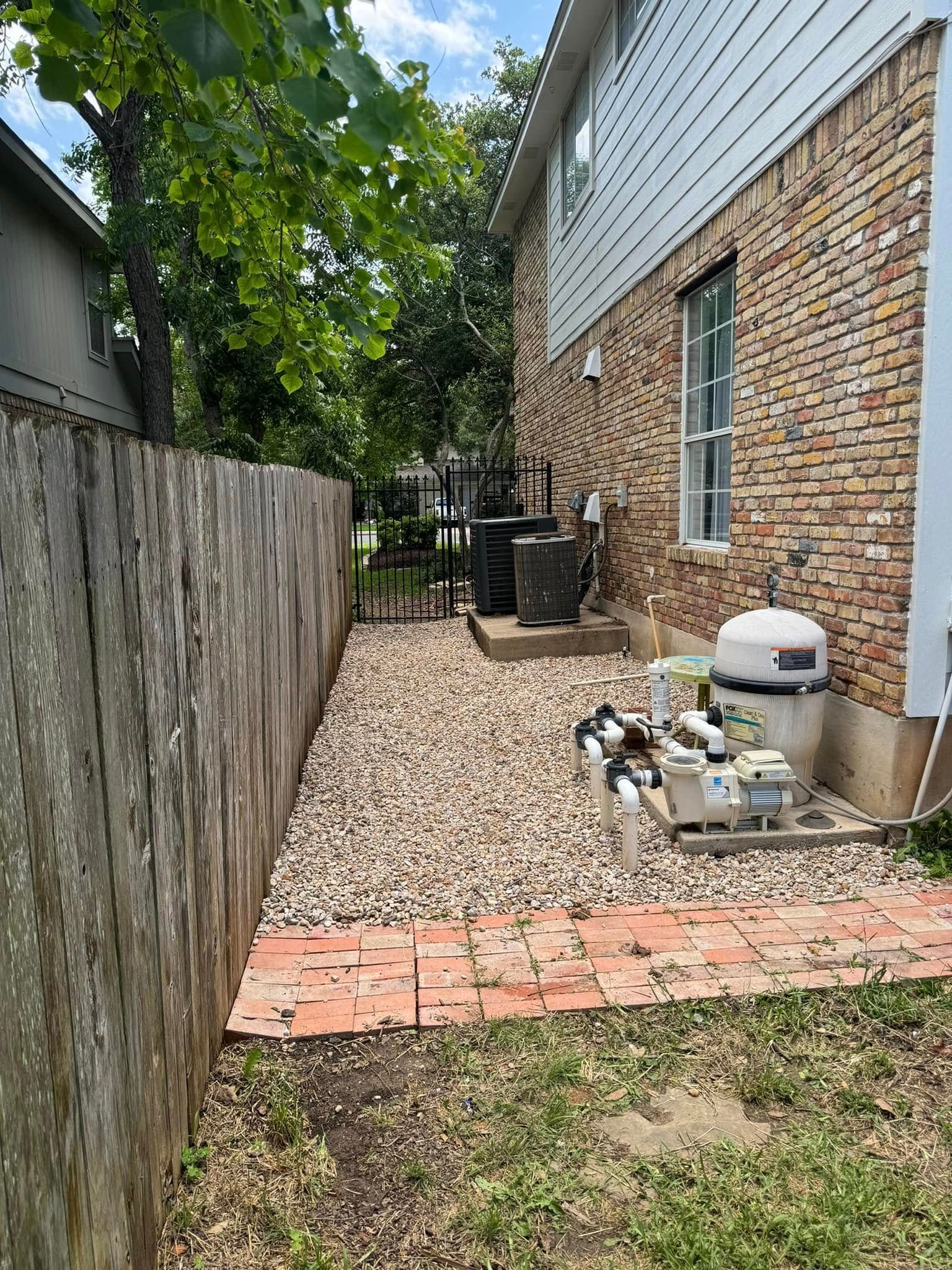 Backyard with a brick house, wooden fence, gravel path, and pool equipment.