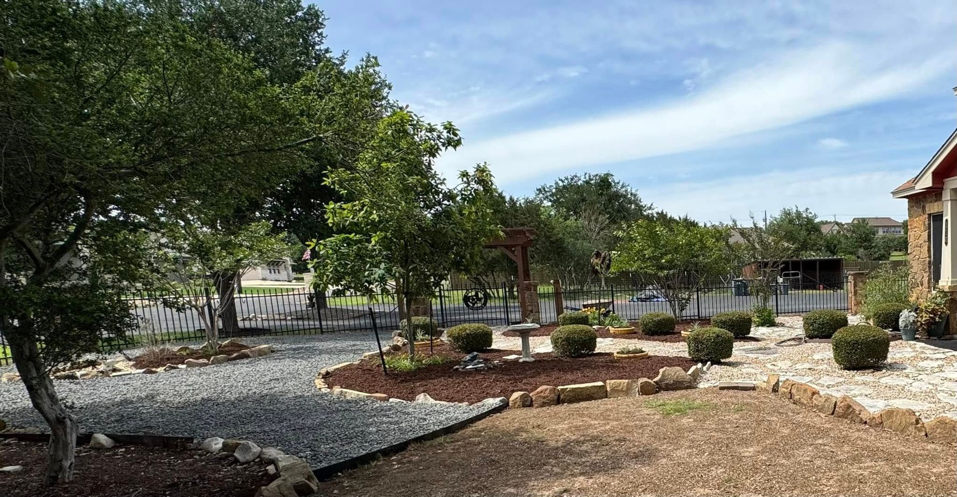 Landscaped yard with trees, gravel, and shrubs, beside a house with a red section, under a partly cloudy sky.