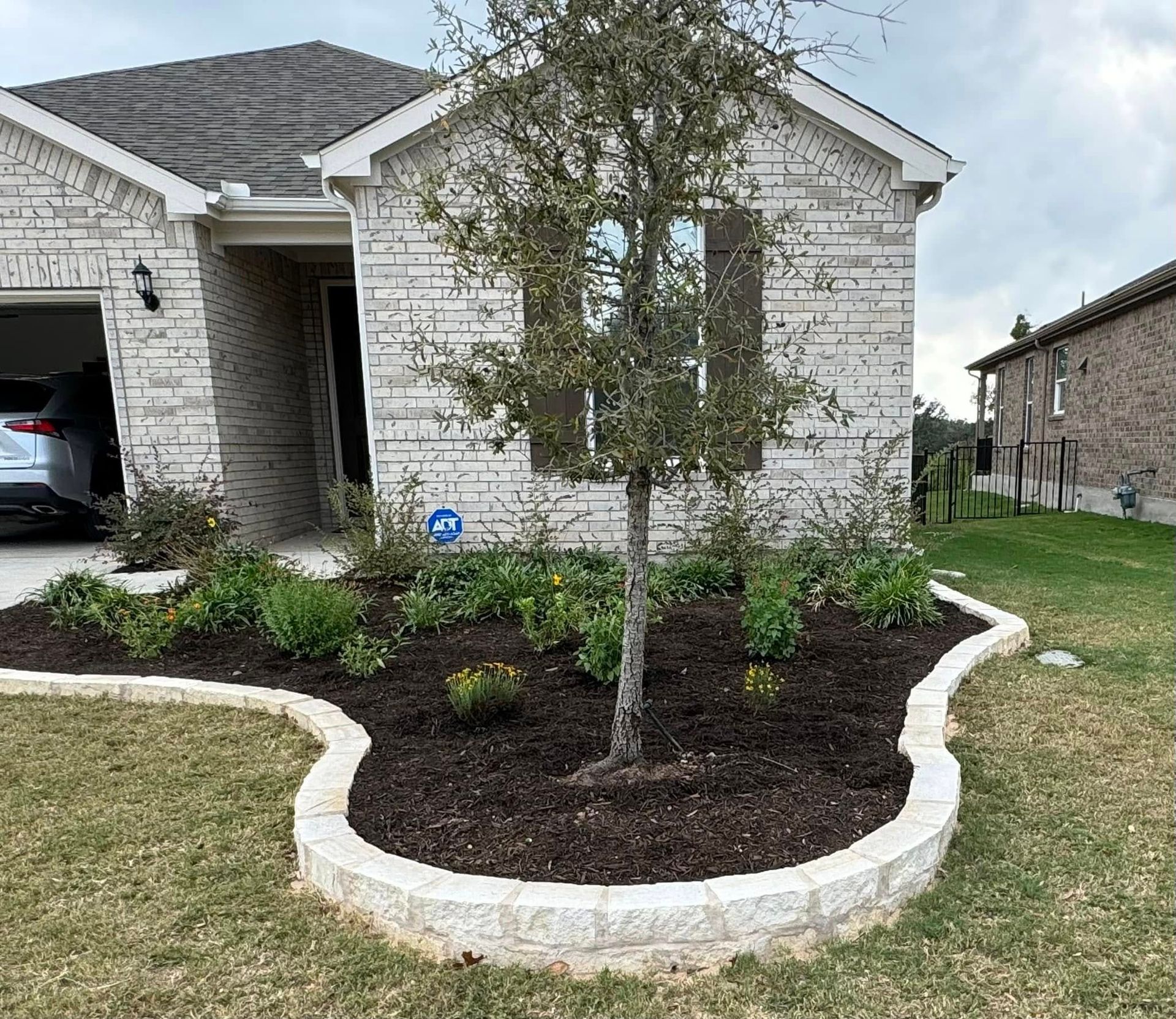 Stone-walled landscaping bed in front of a brick house with a tree and various plants. A car is visible in the garage.