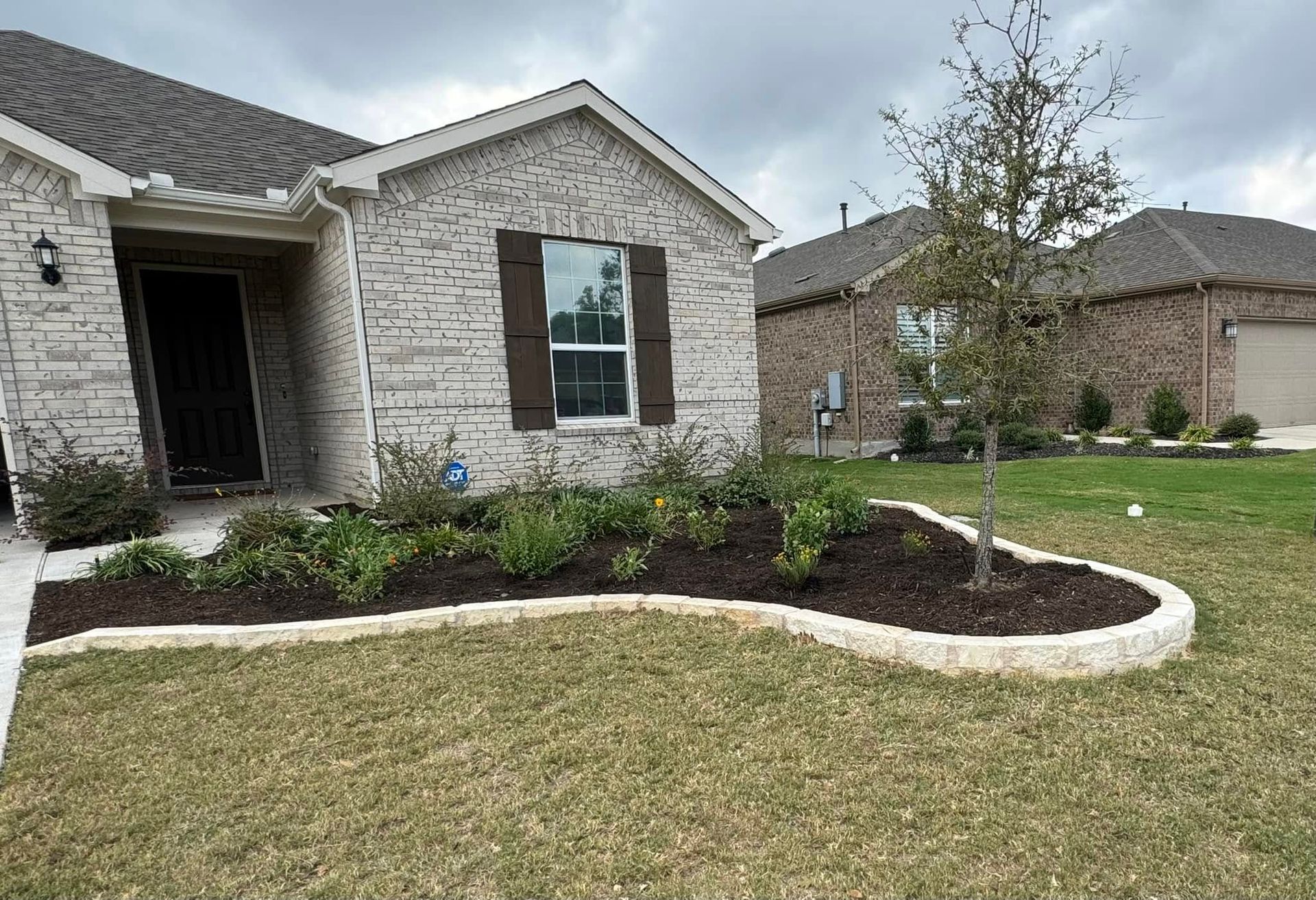 A suburban house with a brick facade and a landscaped front yard with a flower bed, tree, and brown mulch.