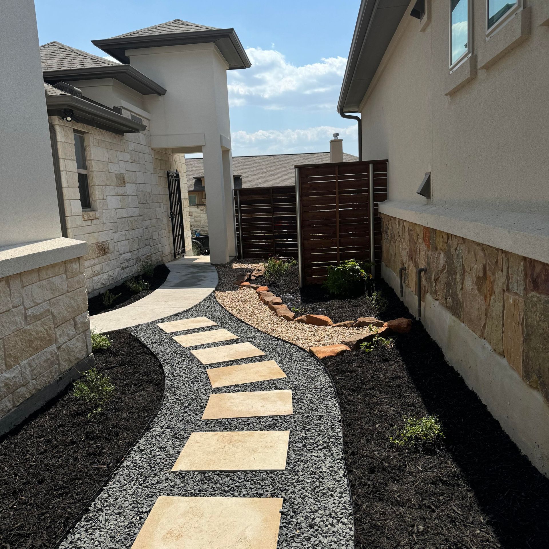 Pathway with stone slabs, leading through a landscaped side yard with dark mulch and gravel, to a dark wood fence.