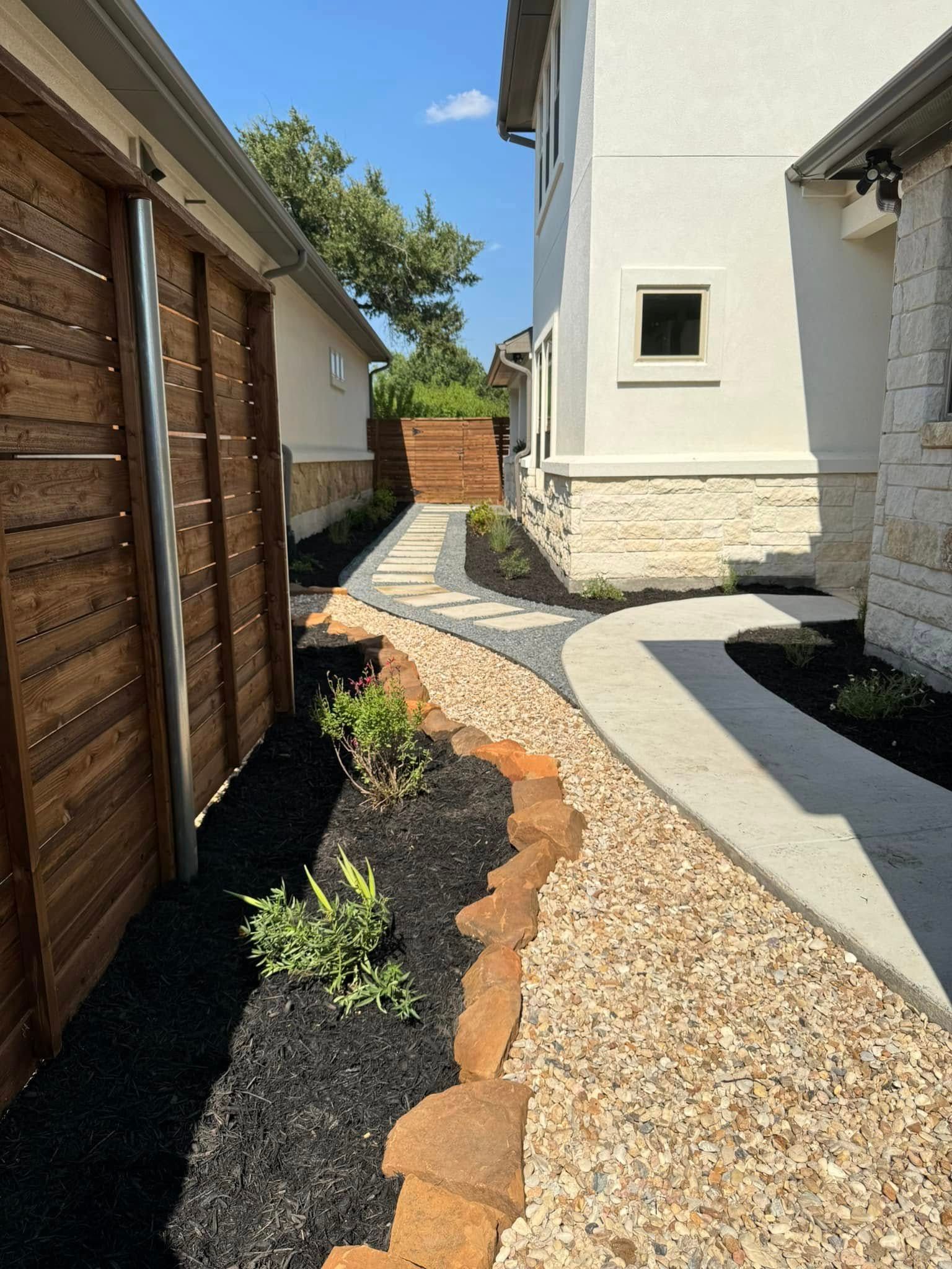Pathway with landscaping between a wooden fence and a white building.