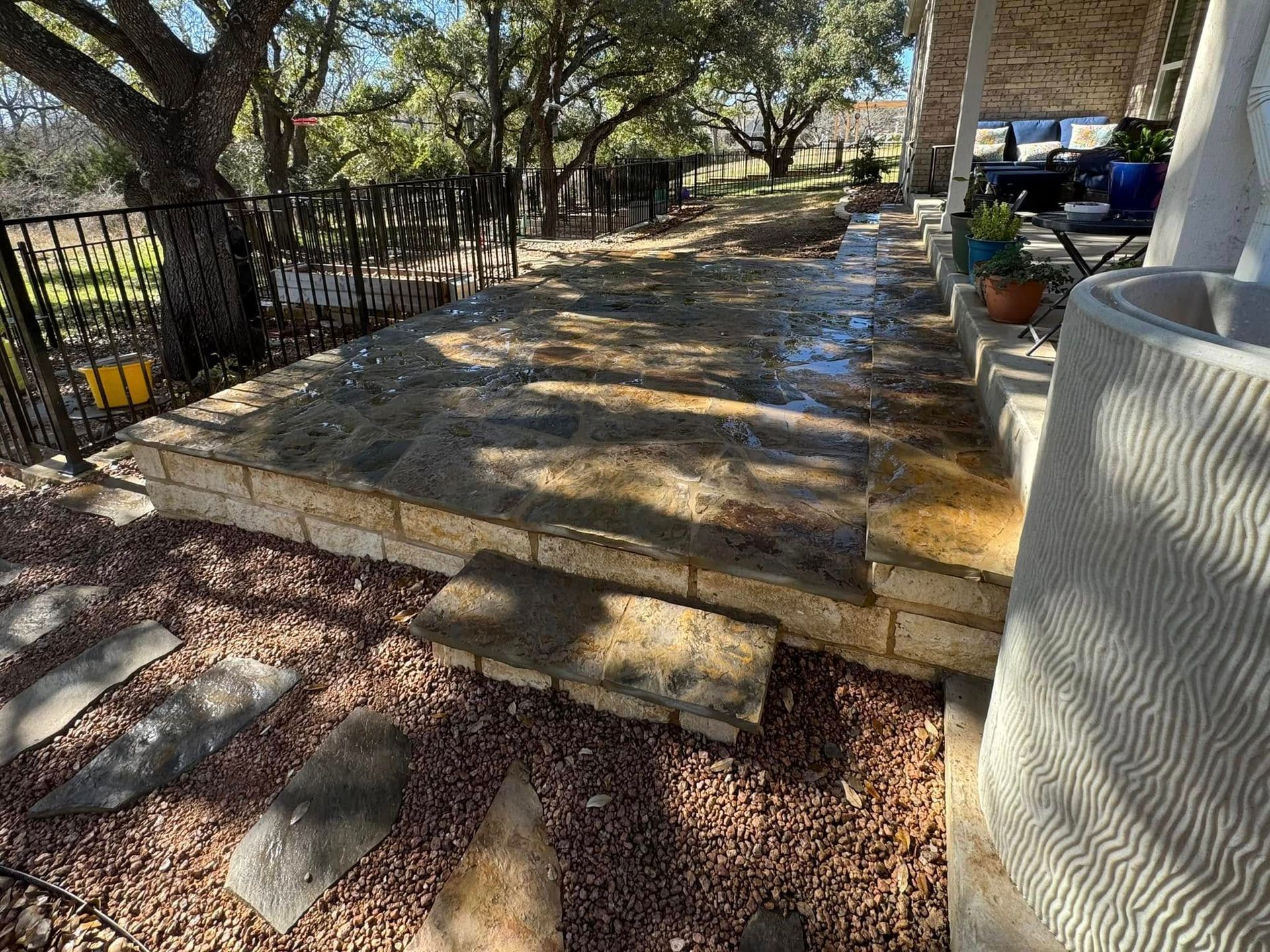 Stone patio and steps with gravel, next to a building and fenced yard, surrounded by trees.