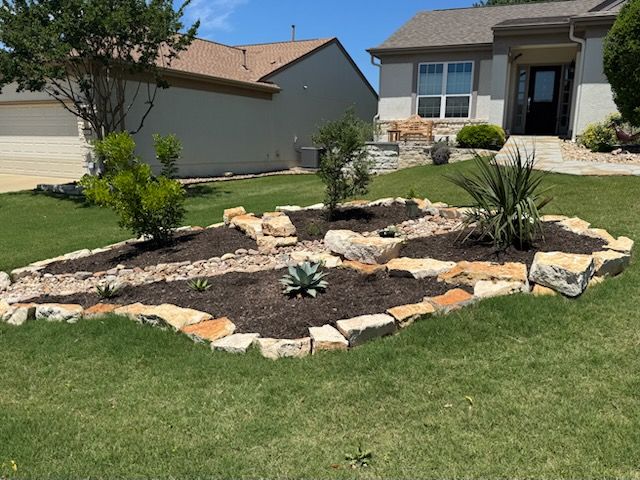 A landscaped flower bed in front of a house. It features rocks, mulch, and various plants. Green lawn surrounds it.