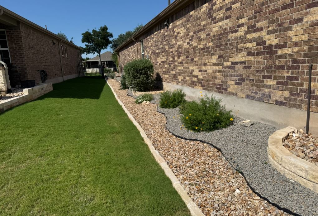 Narrow backyard with grass, brick walls, and landscaped beds with plants and stones.