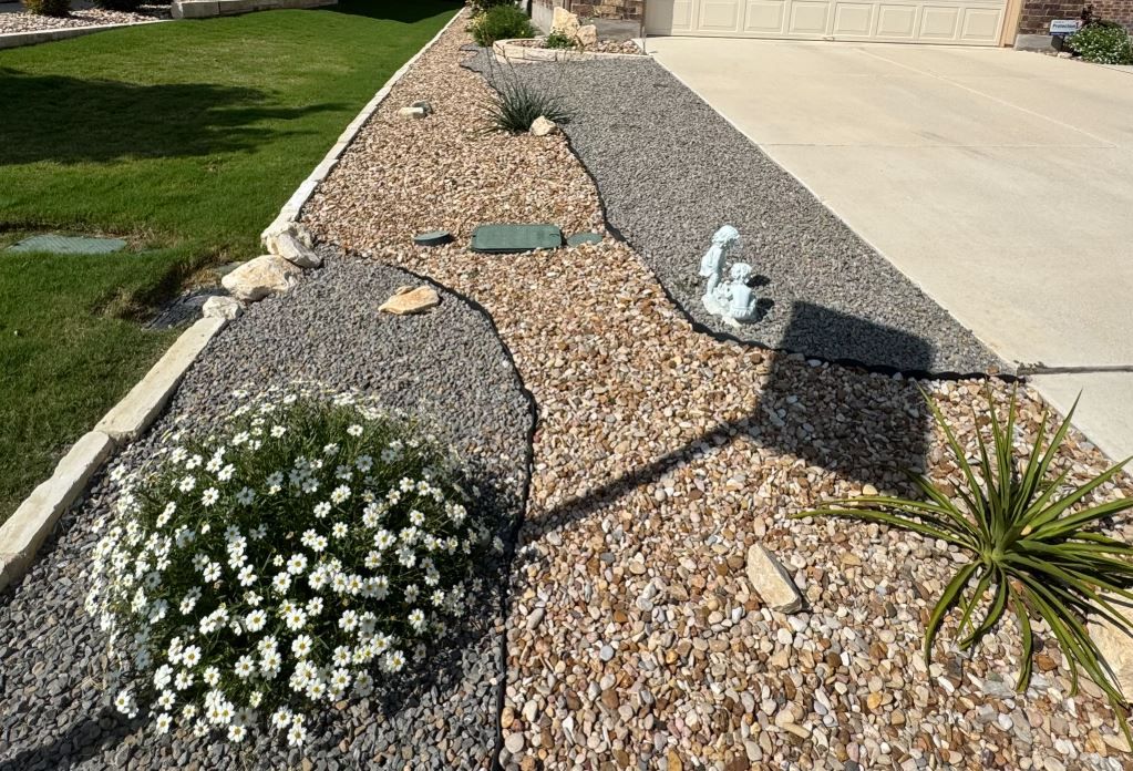 Landscaped rock garden with grey, tan, and light-colored gravel, plants, and driveway.