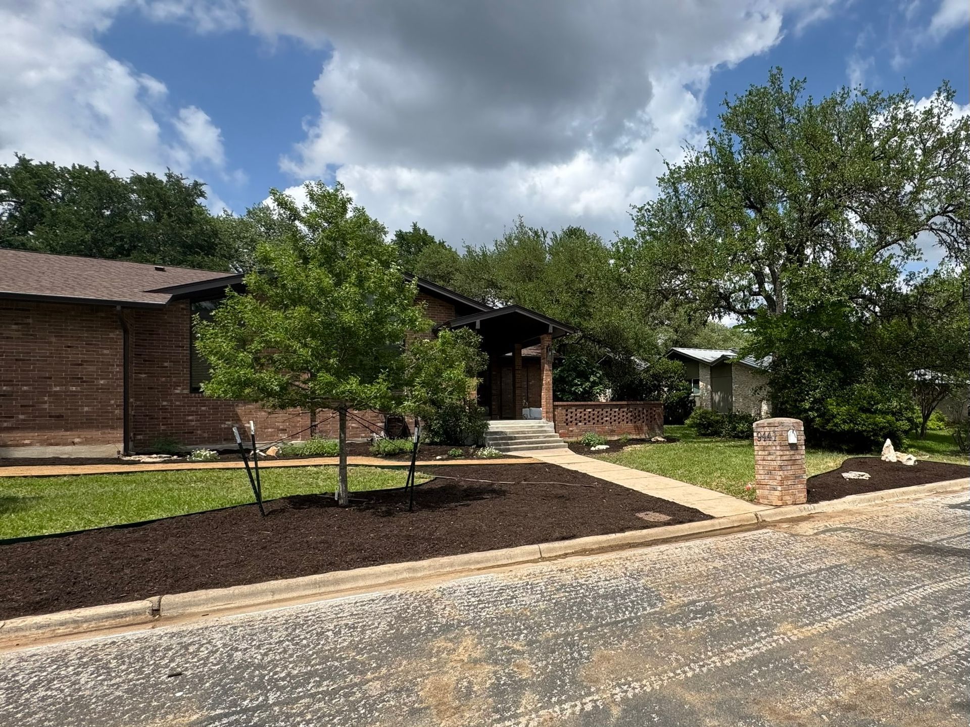 A one-story brick house with a newly mulched yard and a tree-lined walkway under a cloudy sky.