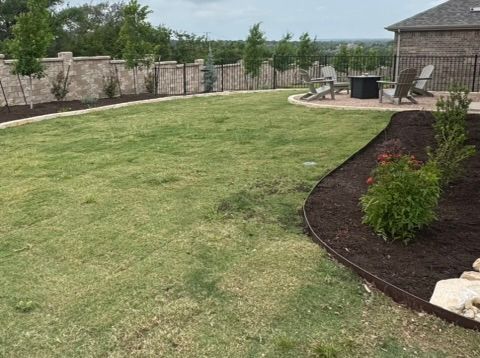 Backyard with green grass, landscaping beds, stone patio with chairs, and stone wall with trees.