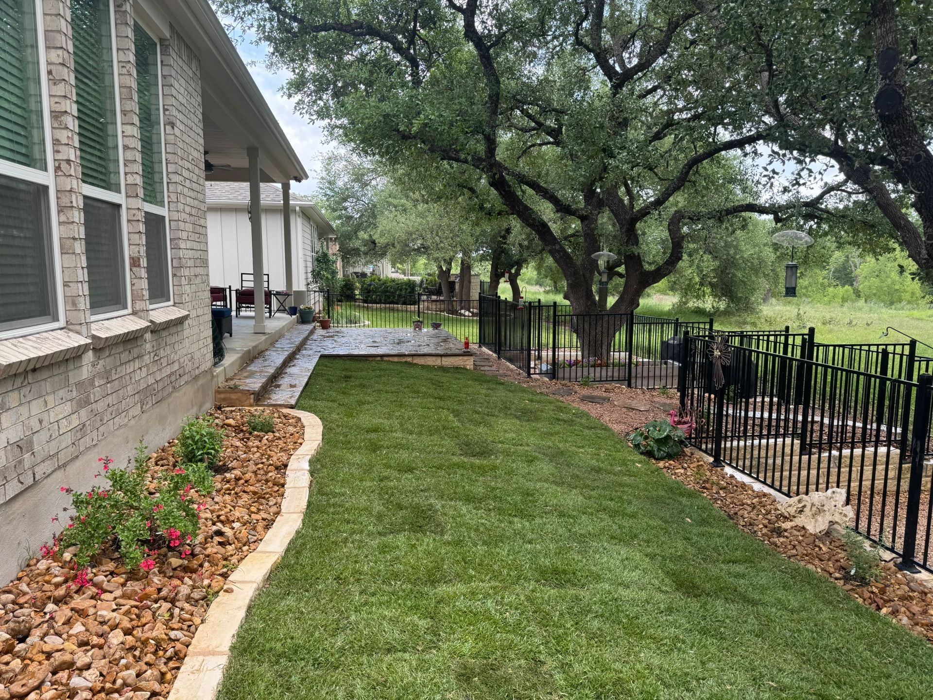 Lush green lawn alongside a house, edged with tan stone and leading to a black fenced area with trees in the background.