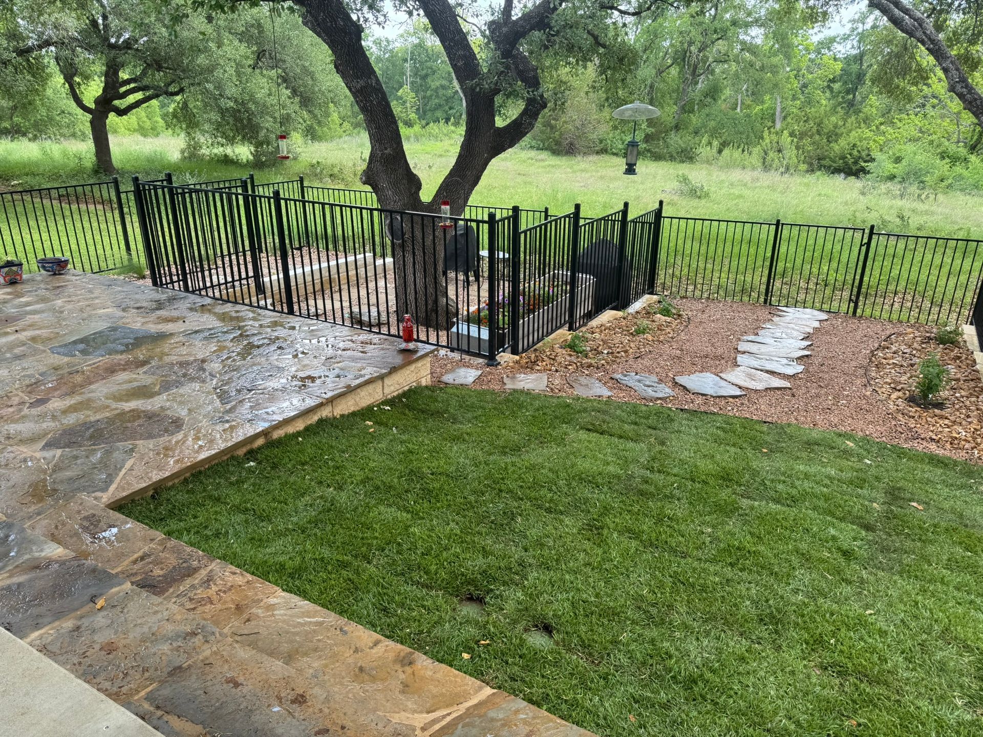 Patio with stone pavers, green lawn, black metal fence, and tree.