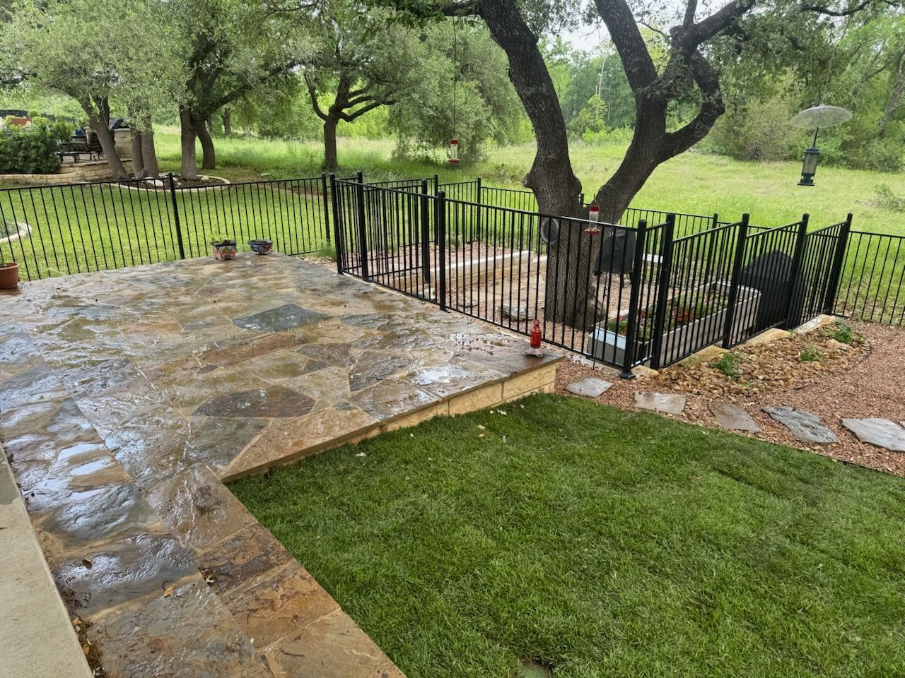 Stone patio and lawn with a black fence, leading to a wooded area, trees in the background.