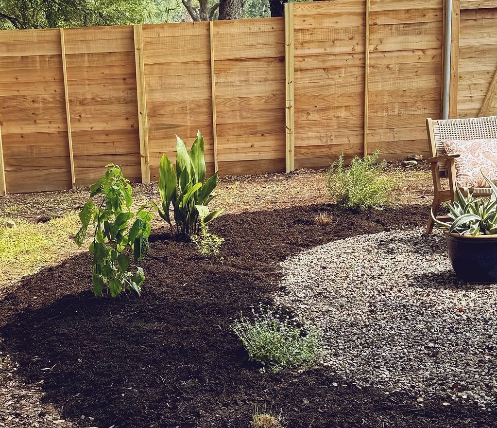 Garden bed with dark mulch and gravel, plants, and a wooden fence.
