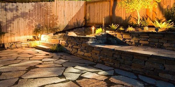 Stone patio and retaining wall illuminated at night, with wooden fence backdrop and small trees.