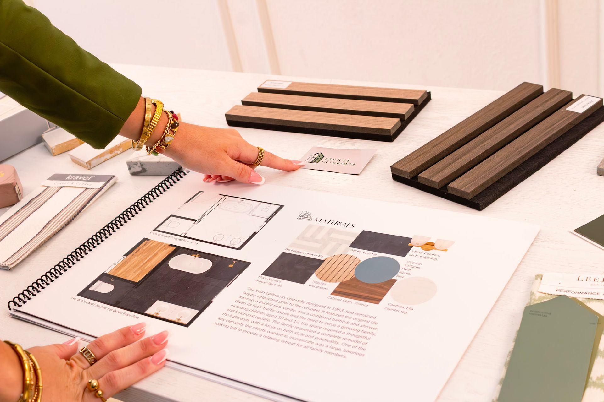 Hands pointing at a design plan and material samples on a table. Gold bracelets and rings are visible.