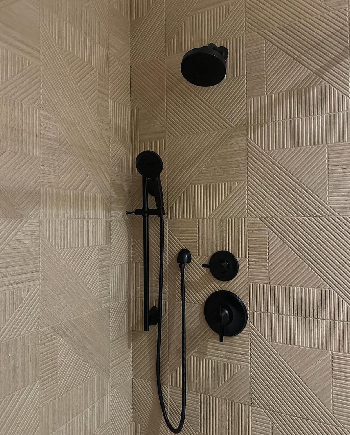 Black shower fixtures on textured beige tile wall. Includes showerhead, handheld, and controls.