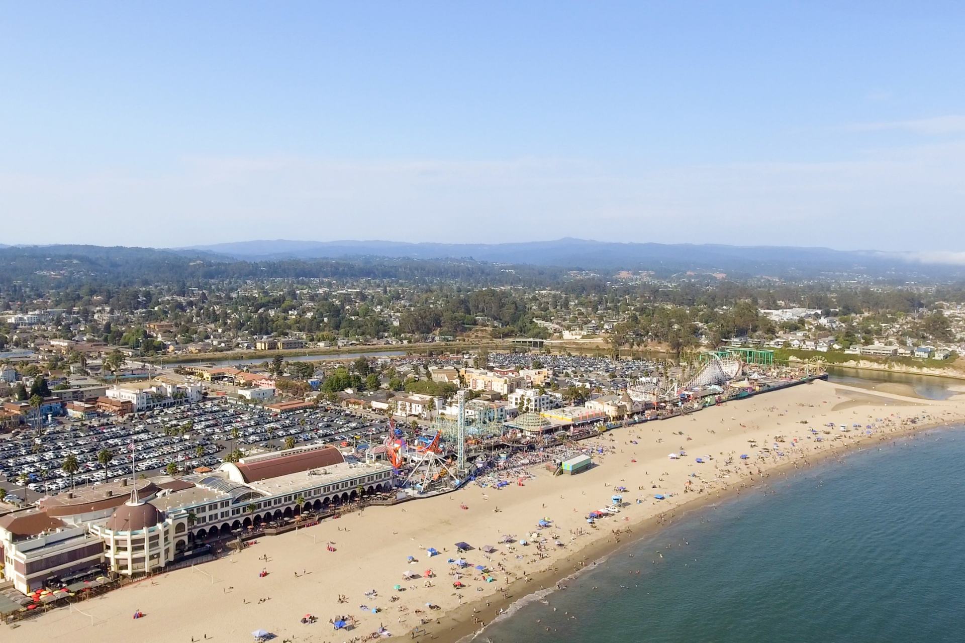 Aerial view of a crowded sandy beach with blue ocean, shoreline buildings, and a distant cityscape.