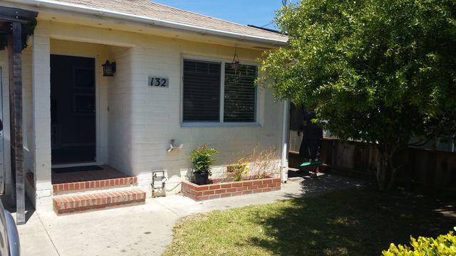 Front of a white house with a porch, dark door, window shutters, and a tree shading the yard.