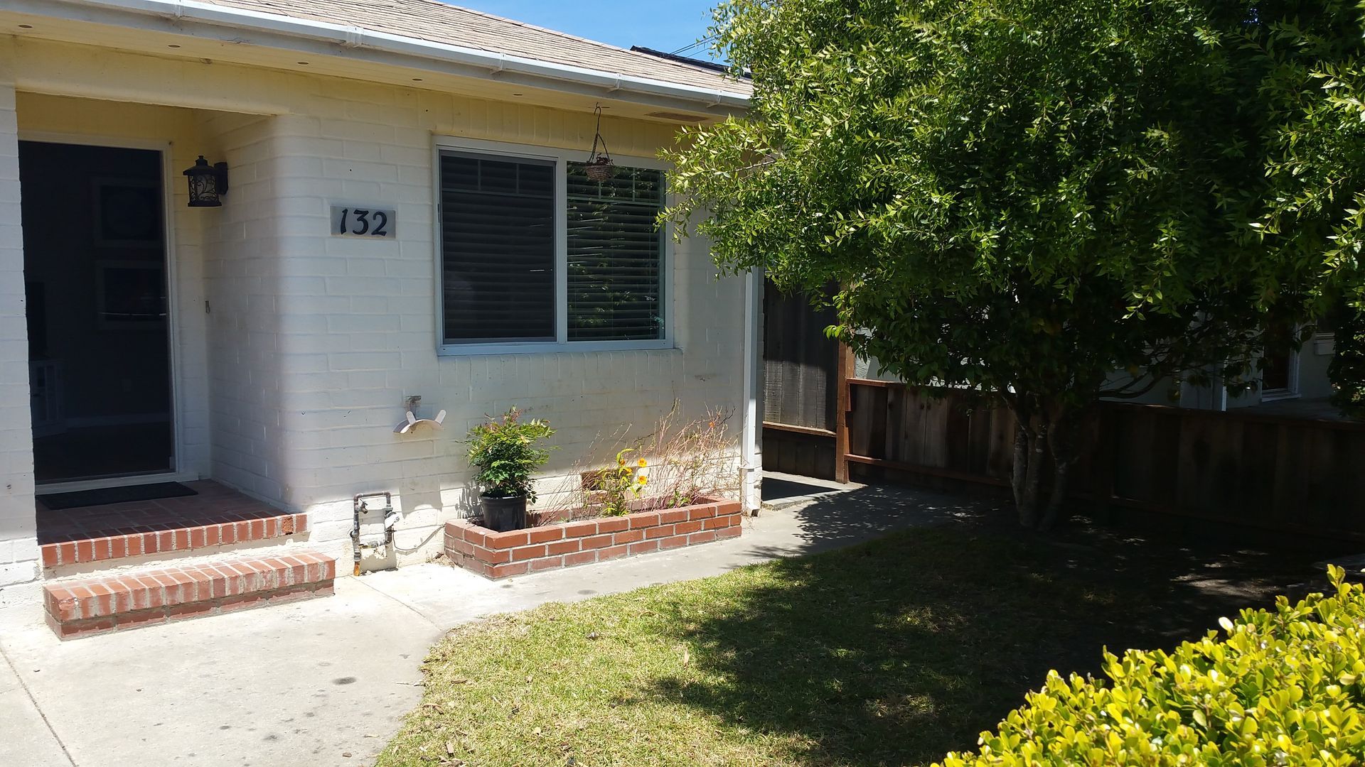 Sunlit front porch of a white house with black shutters, brick steps, and a shaded front yard