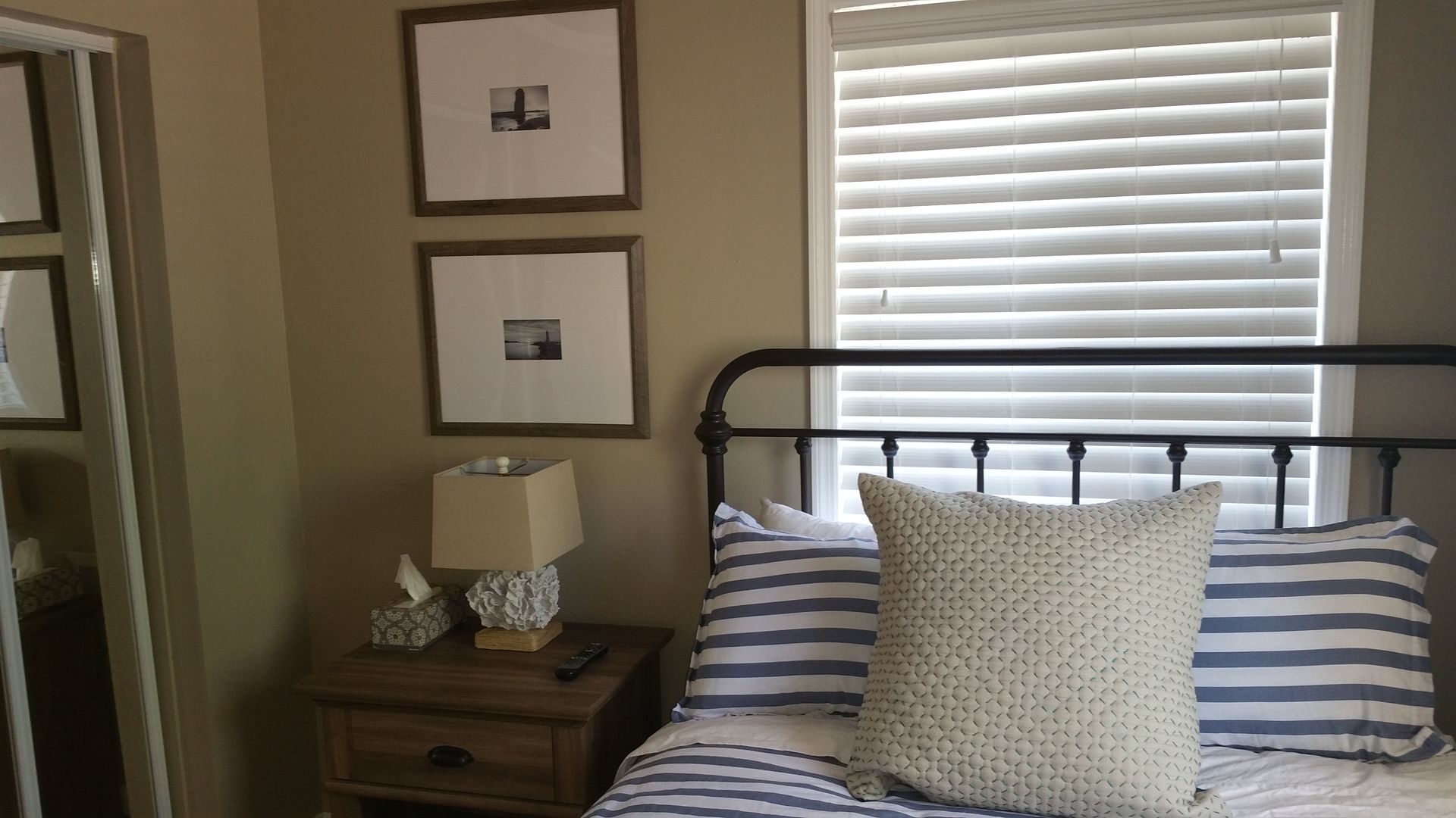 Bedroom with striped bedspread, white pillow, bedside lamp, framed art, and window blinds