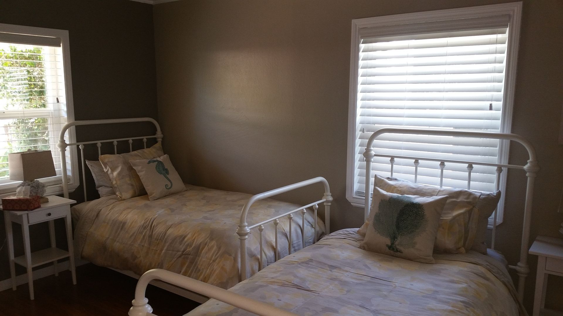 Bedroom with two twin beds, white metal frames, a window with blinds, and a small side table by a sunlit window