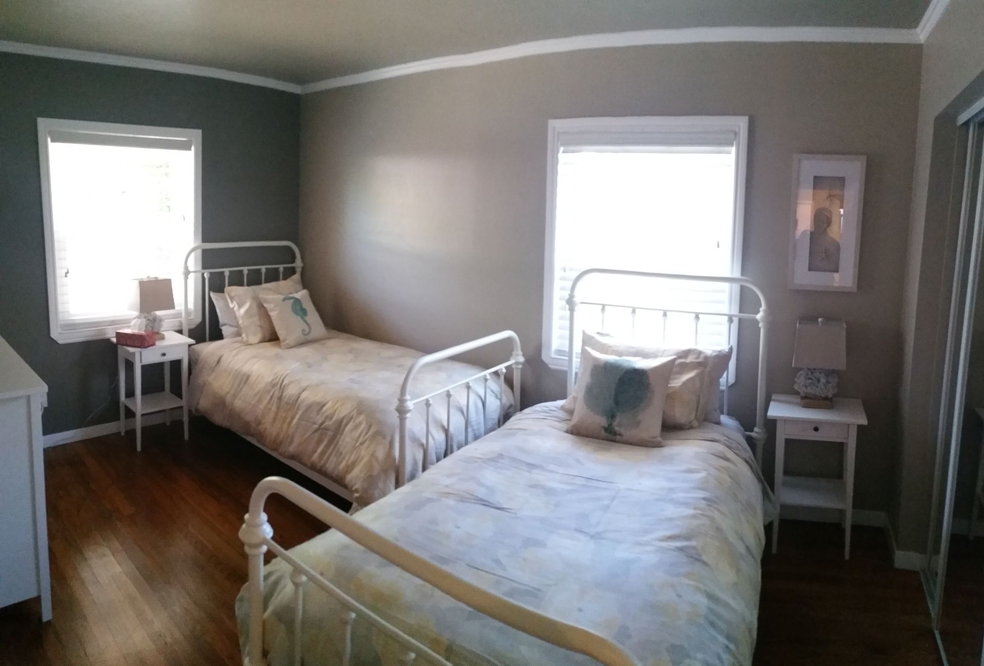Bright bedroom with two white metal beds, gray walls, hardwood floor, and two windows