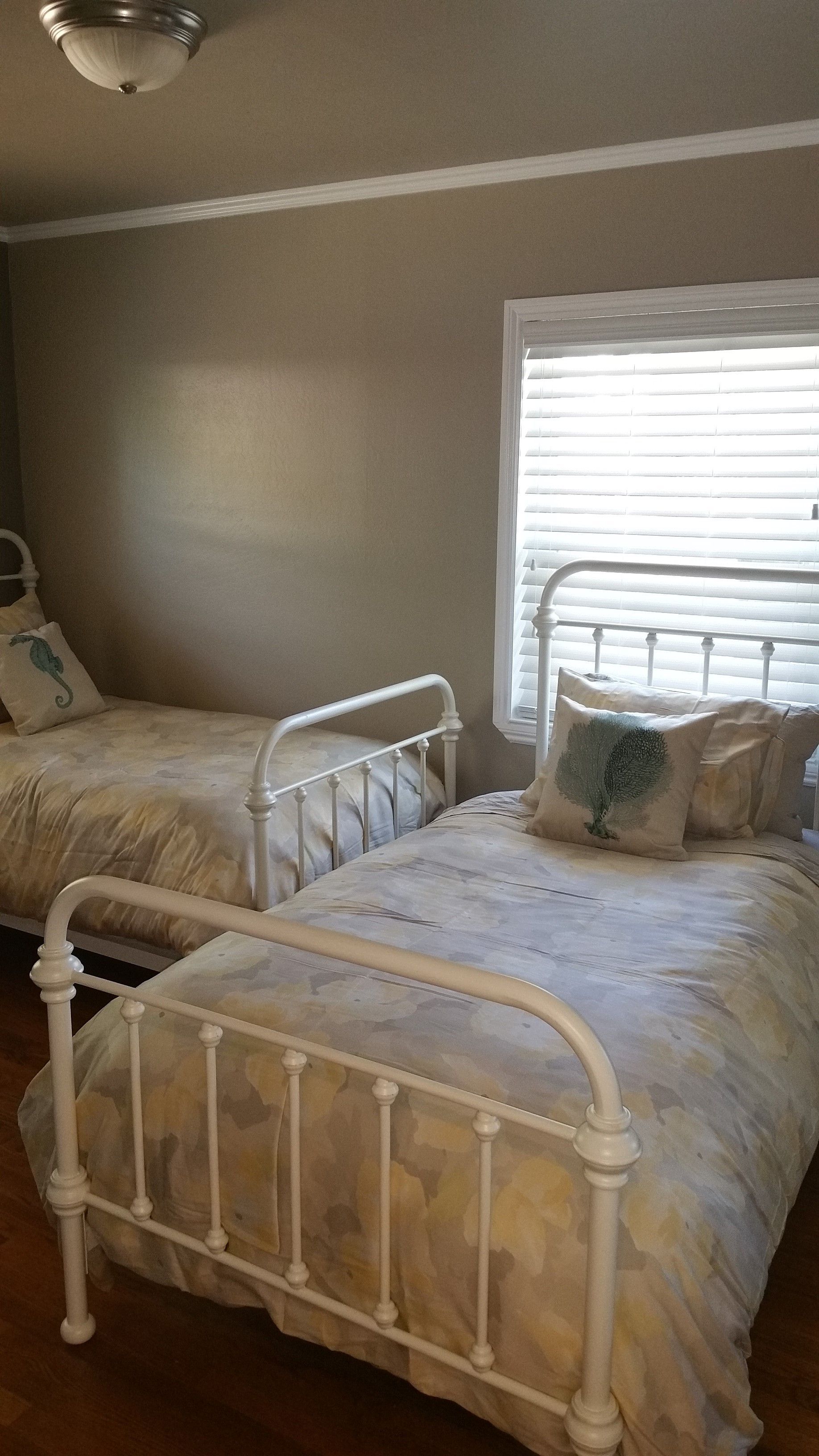 Small bedroom with two white metal twin beds, beige walls, and a window with blinds.