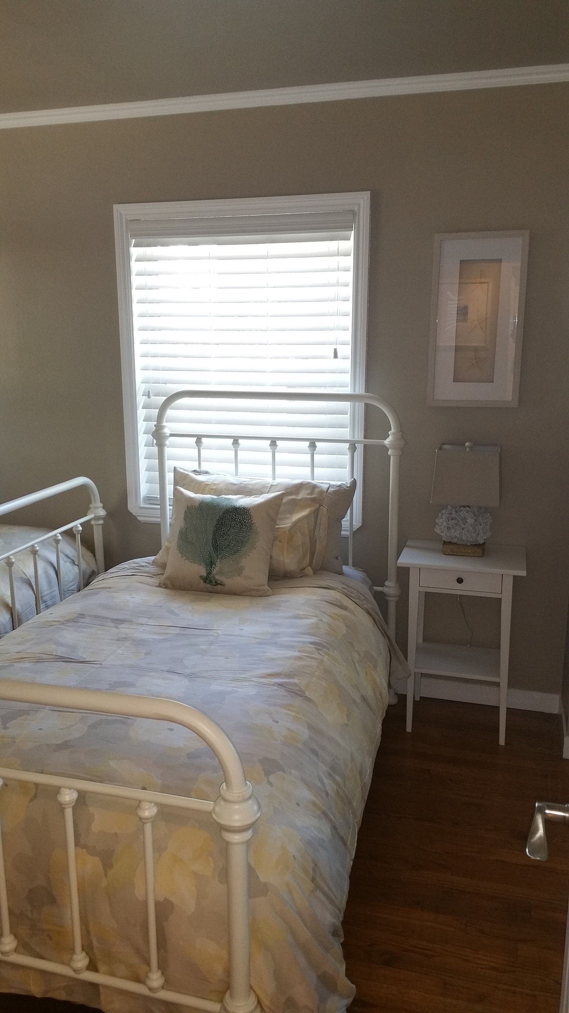 Bright bedroom with a white metal bed, patterned bedding, window blinds, and a small bedside table.