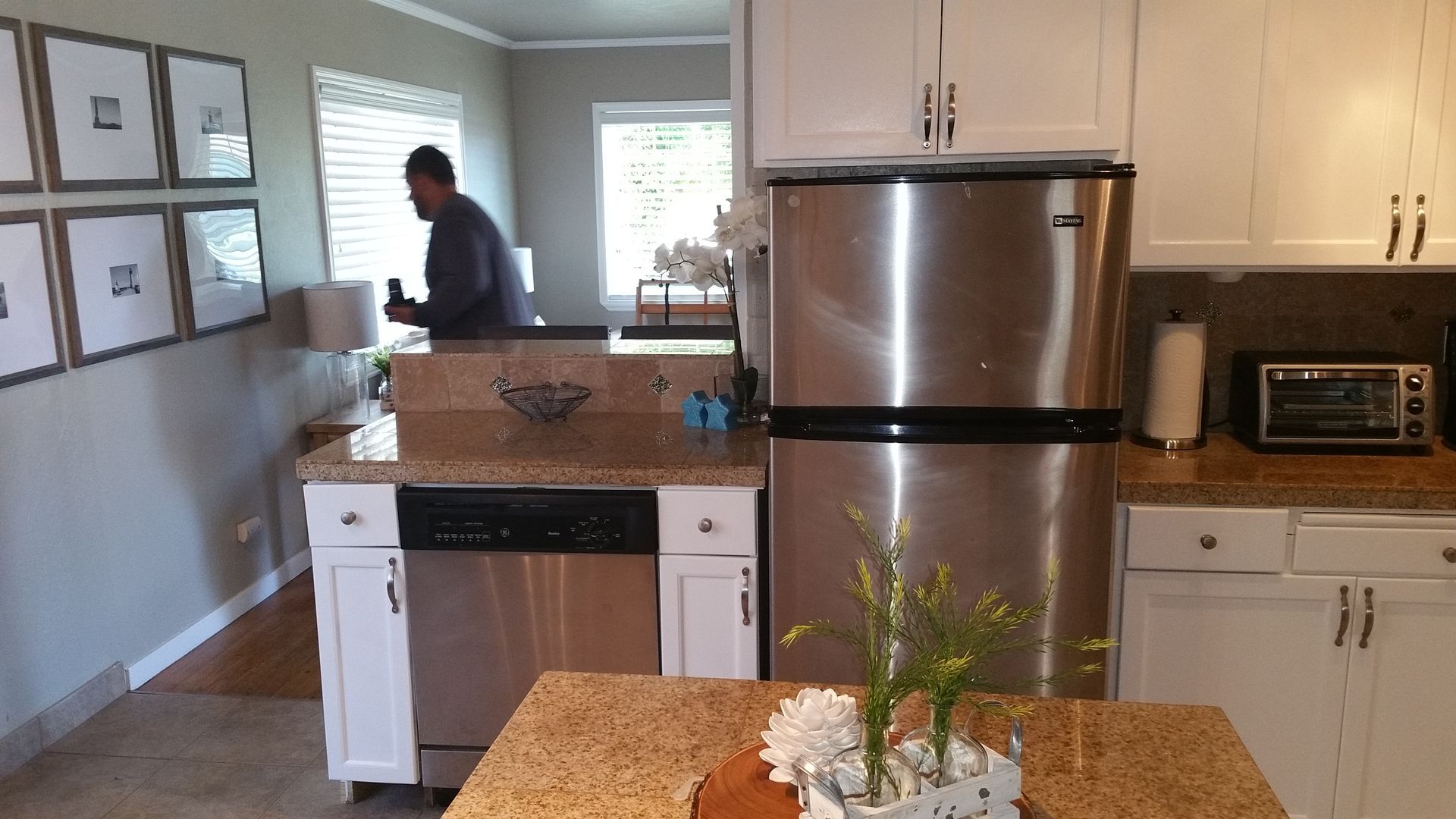 Bright kitchen with stainless steel fridge, white cabinets, granite island, and a person standing near the back window.