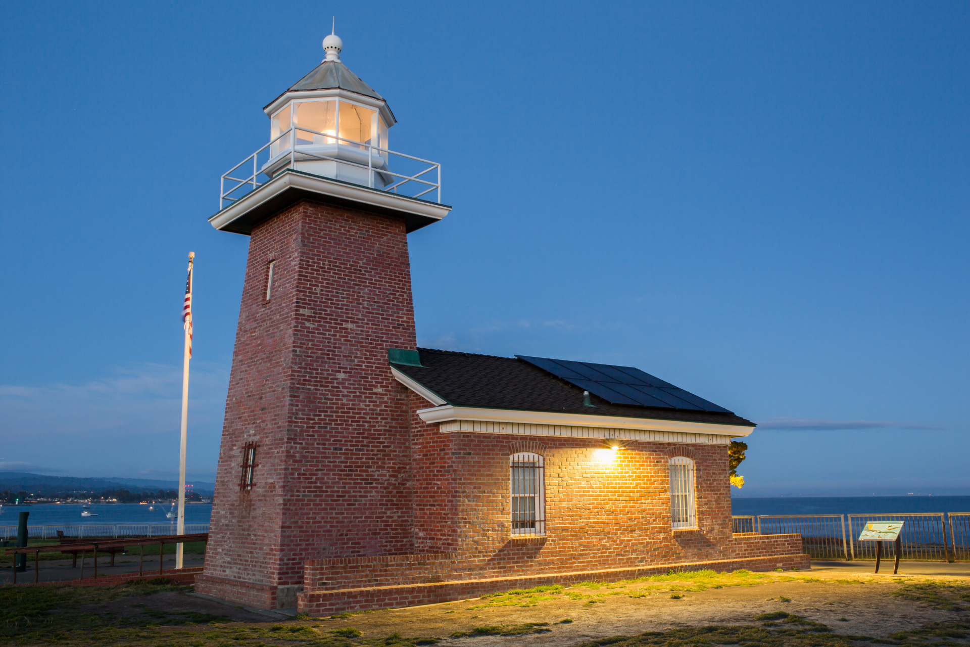 Brick lighthouse at dusk by the water, with a lit lantern room and a small attached building.