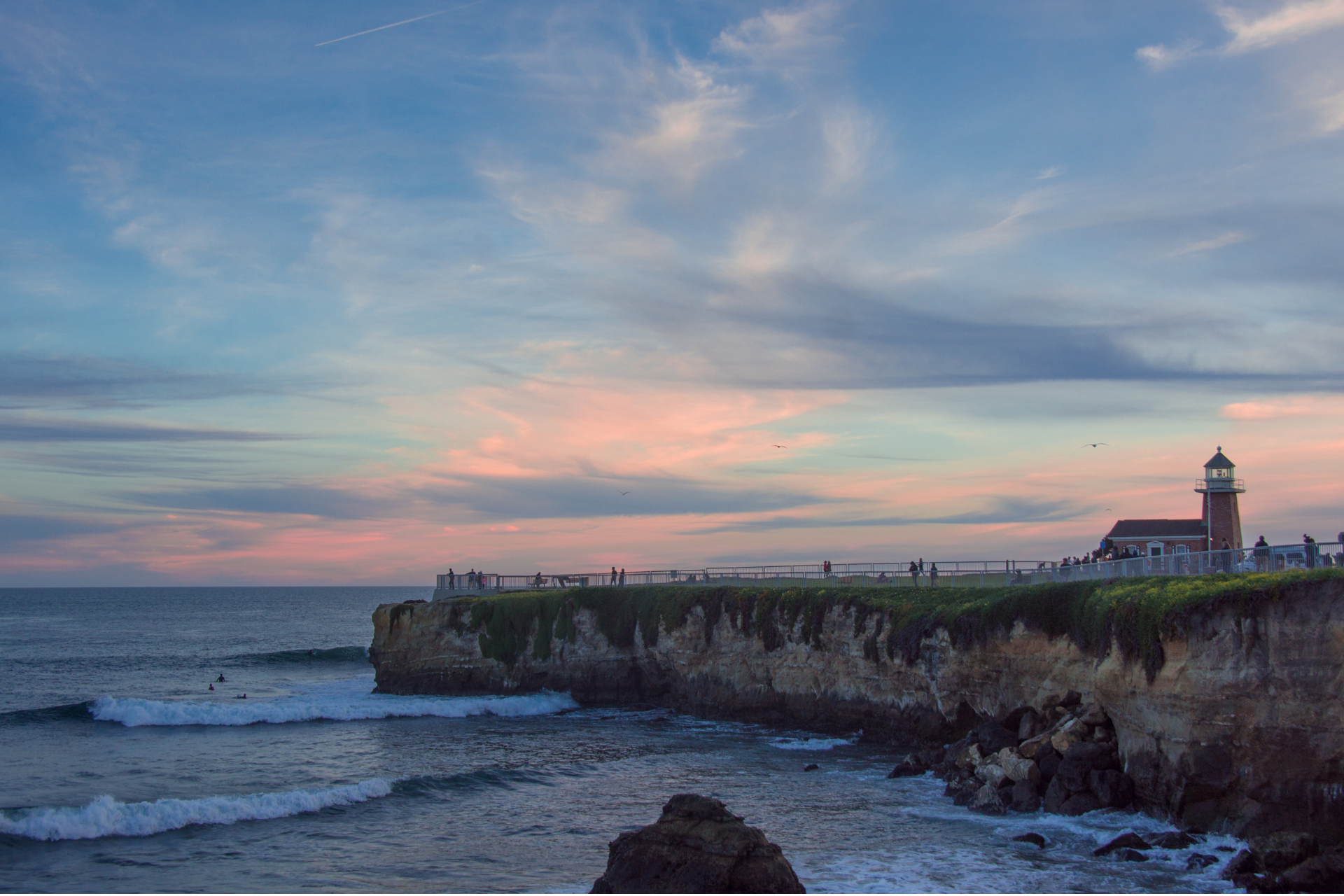Coastal cliff at sunset with waves, pink sky, and a lighthouse on the headland
