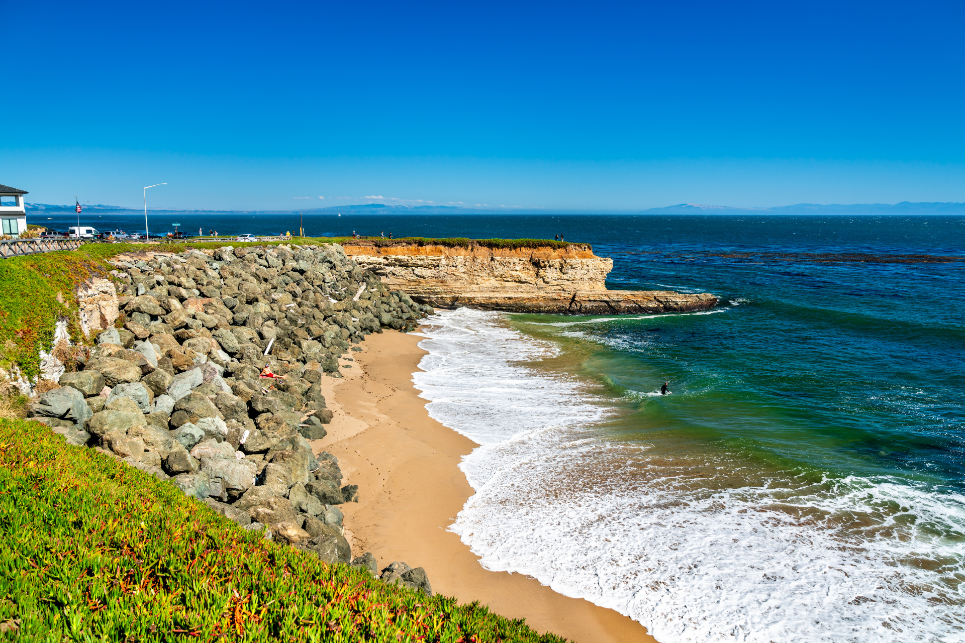 Sunny coastal beach with waves, rocky cliff, and blue ocean under a clear sky