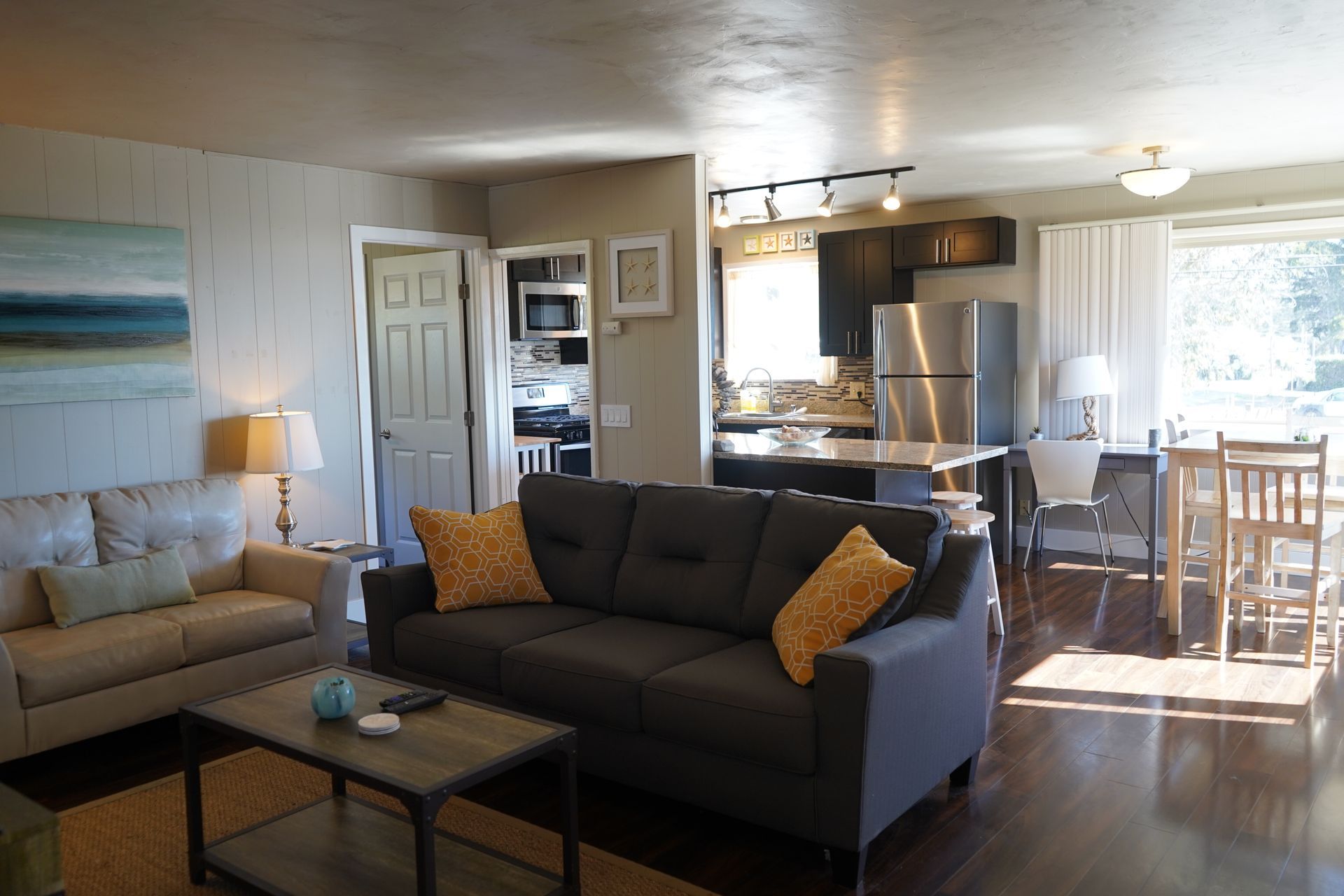 Open-plan living room with gray sofa, coffee table, dining area, and bright kitchen by a sunlit window