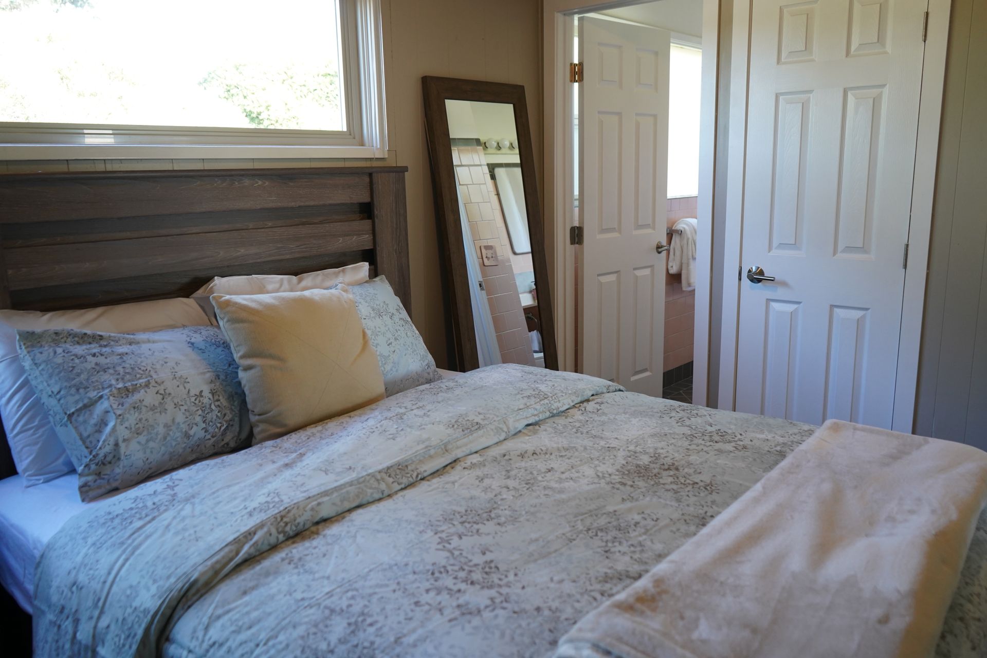 Cozy bedroom with a neatly made bed, pillows, mirror, and closet doors beside a window