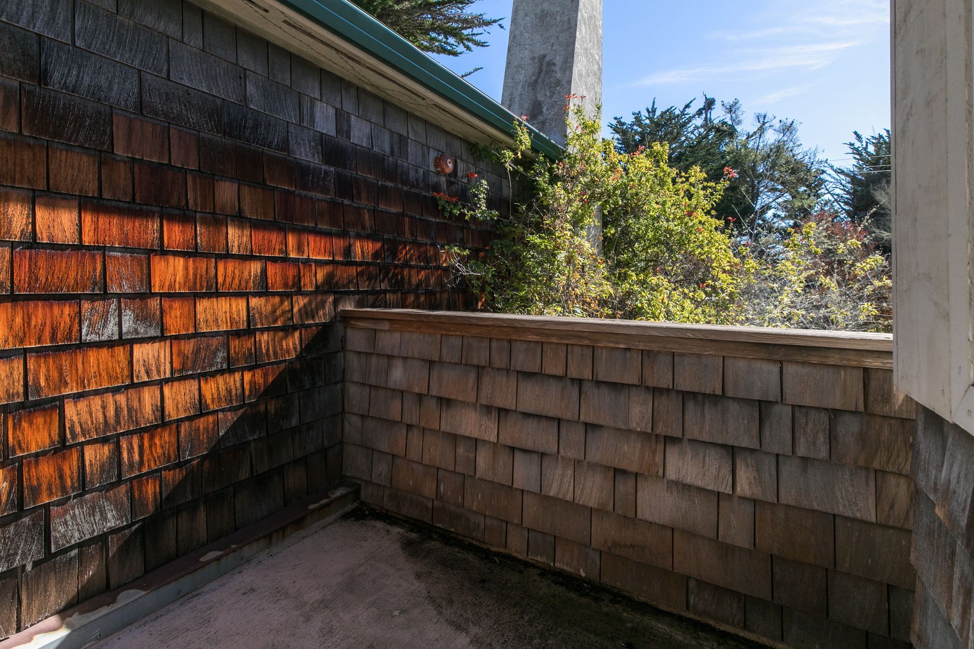 Sunlit wooden shingle wall and low fence on a shaded patio, with trees beyond