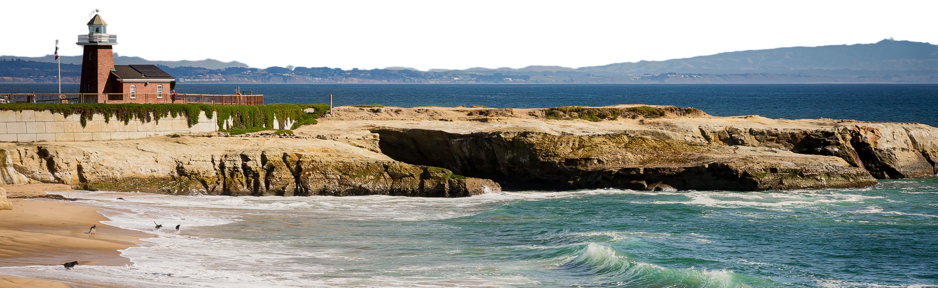 Rocky coastline with a red lighthouse beside calm blue water and sandy shore under a clear sky