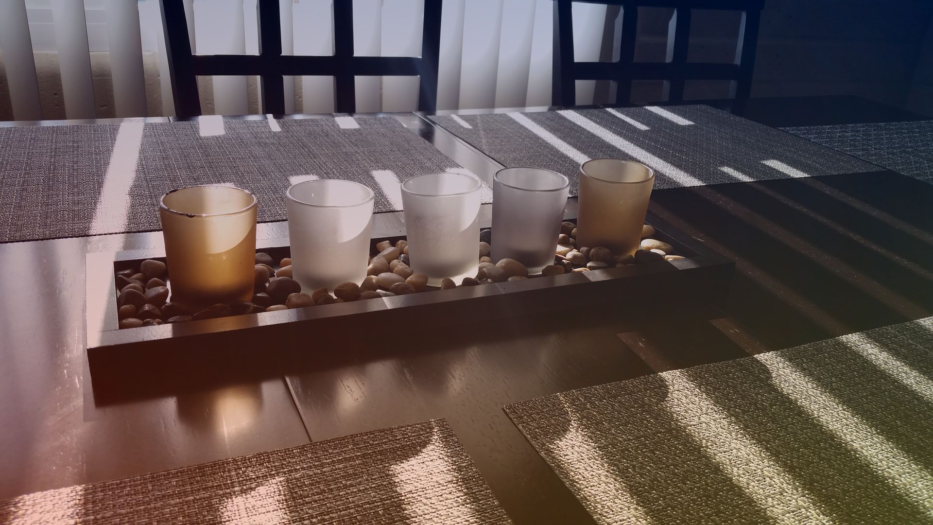 Five small candles in glass holders on a table, with chairs in the background and sunlight casting shadows.