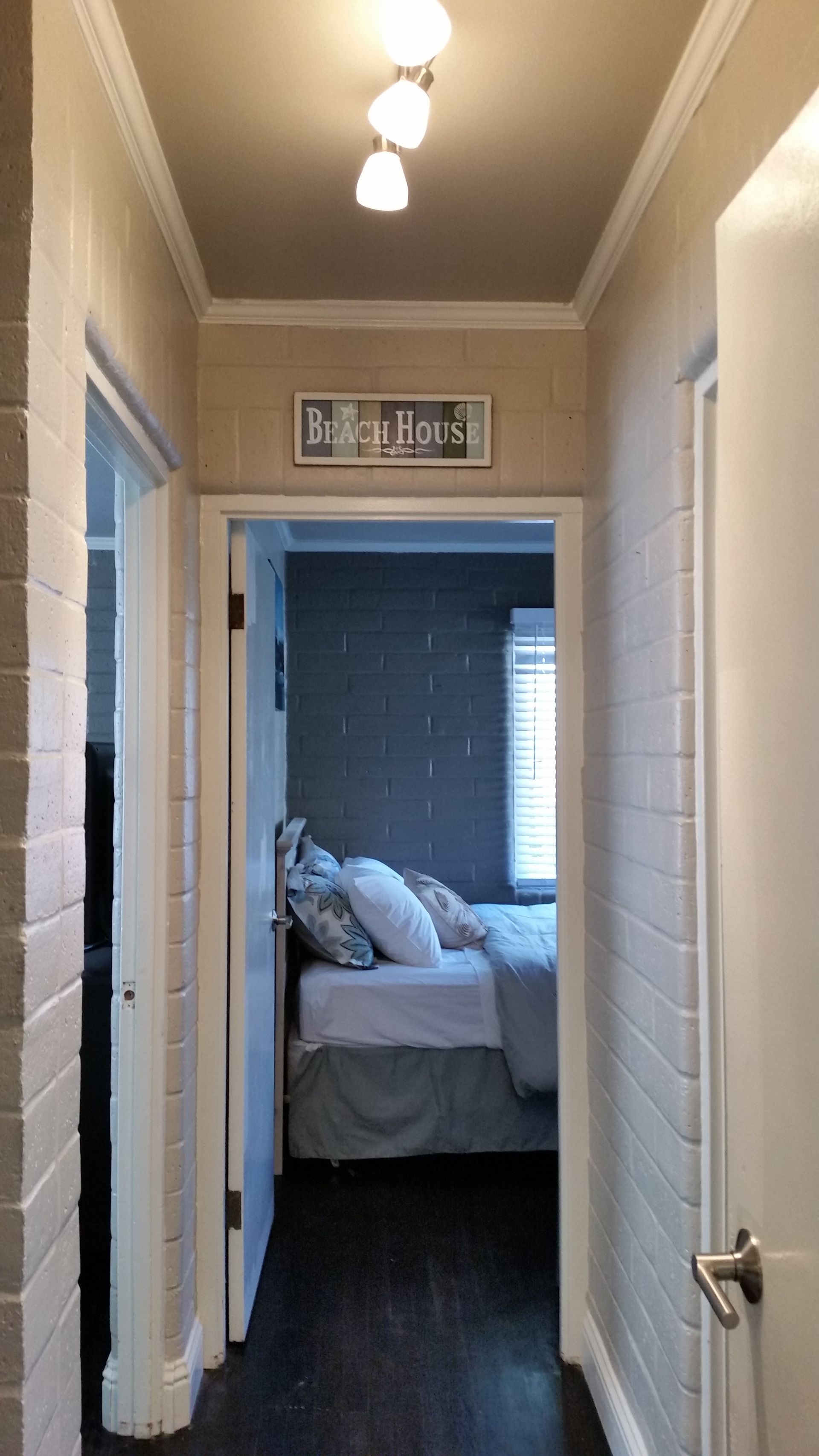 Narrow hallway with white walls and ceiling lights, opening into a bedroom with a made bed and window.