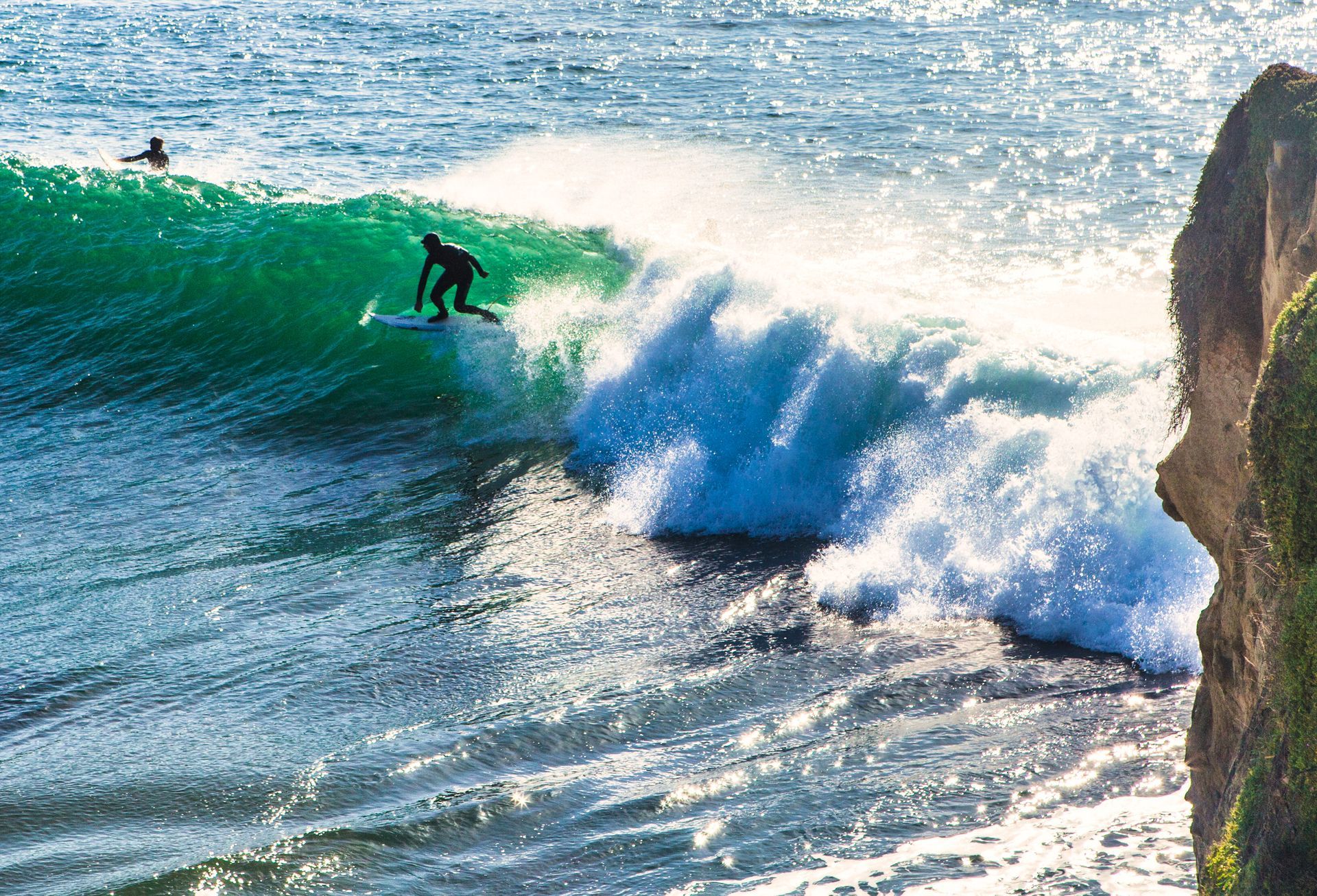 Surfer riding a green wave beside a rocky cliff, with white spray in the sunlight