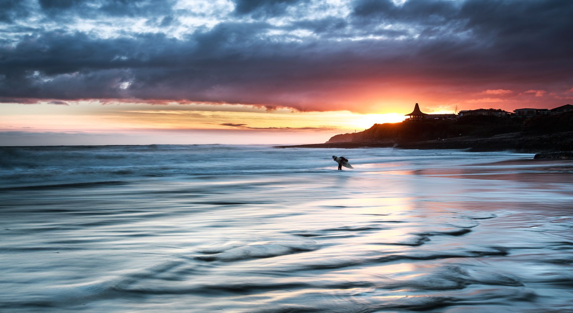 Surfer riding a wave at sunset, with dramatic clouds over a rocky coastline and glowing horizon.