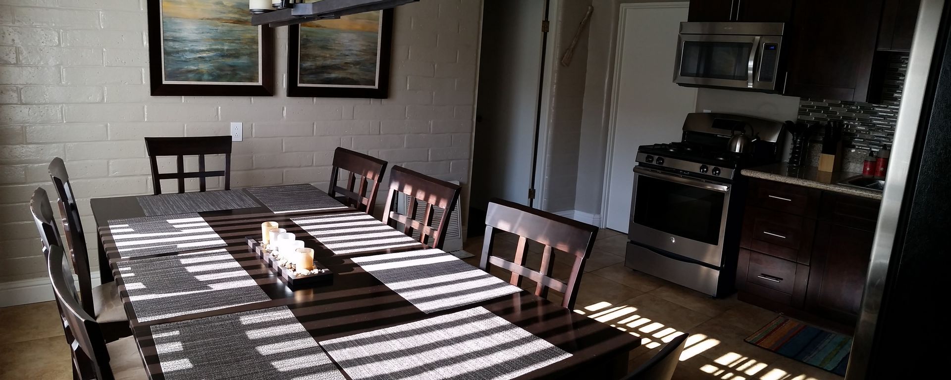 Dining room with a long table, chairs, and sunlight streaming through the windows onto the floor.