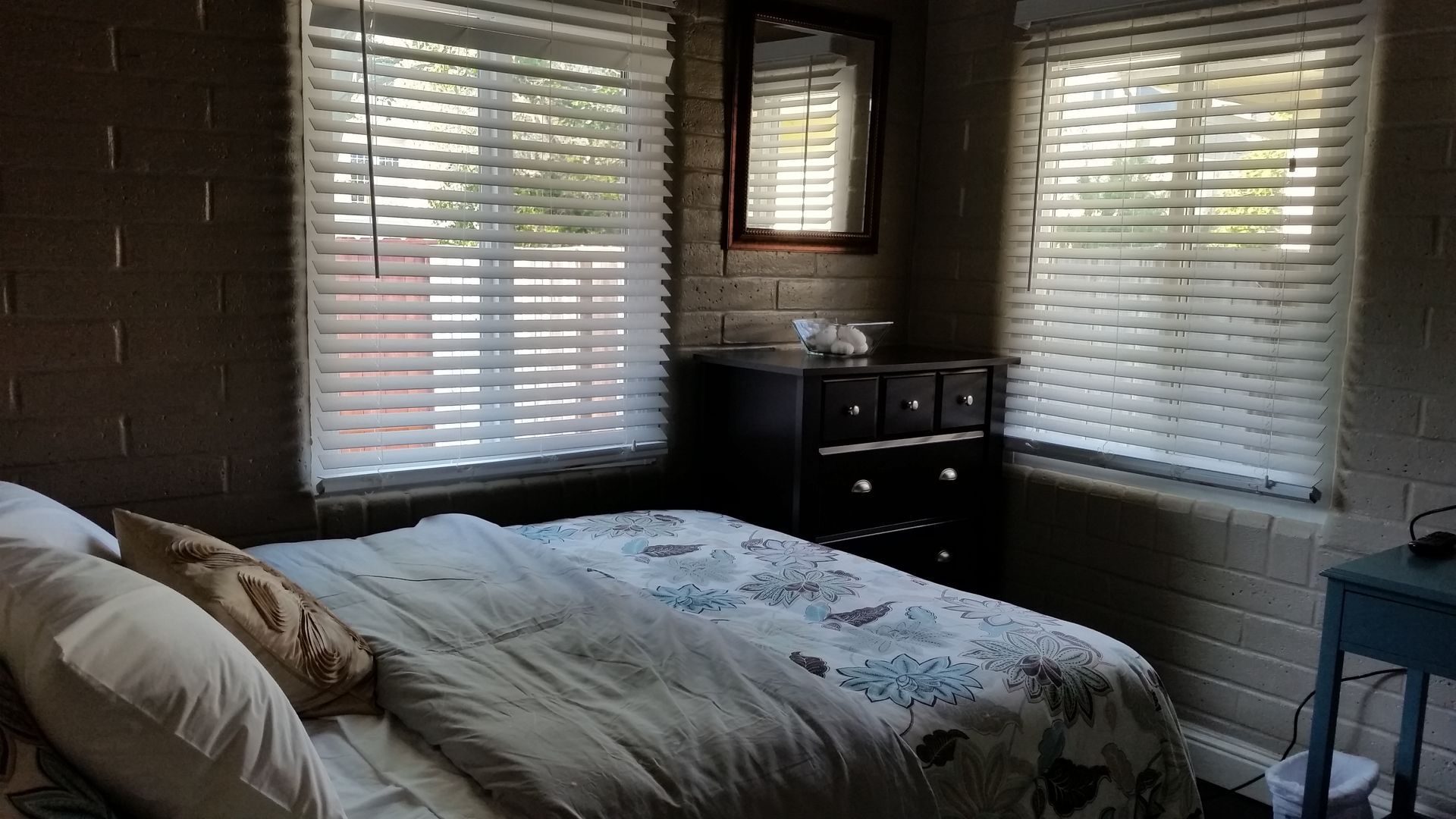 Bedroom with a bed, dark dresser, and two windows with blinds letting in daylight