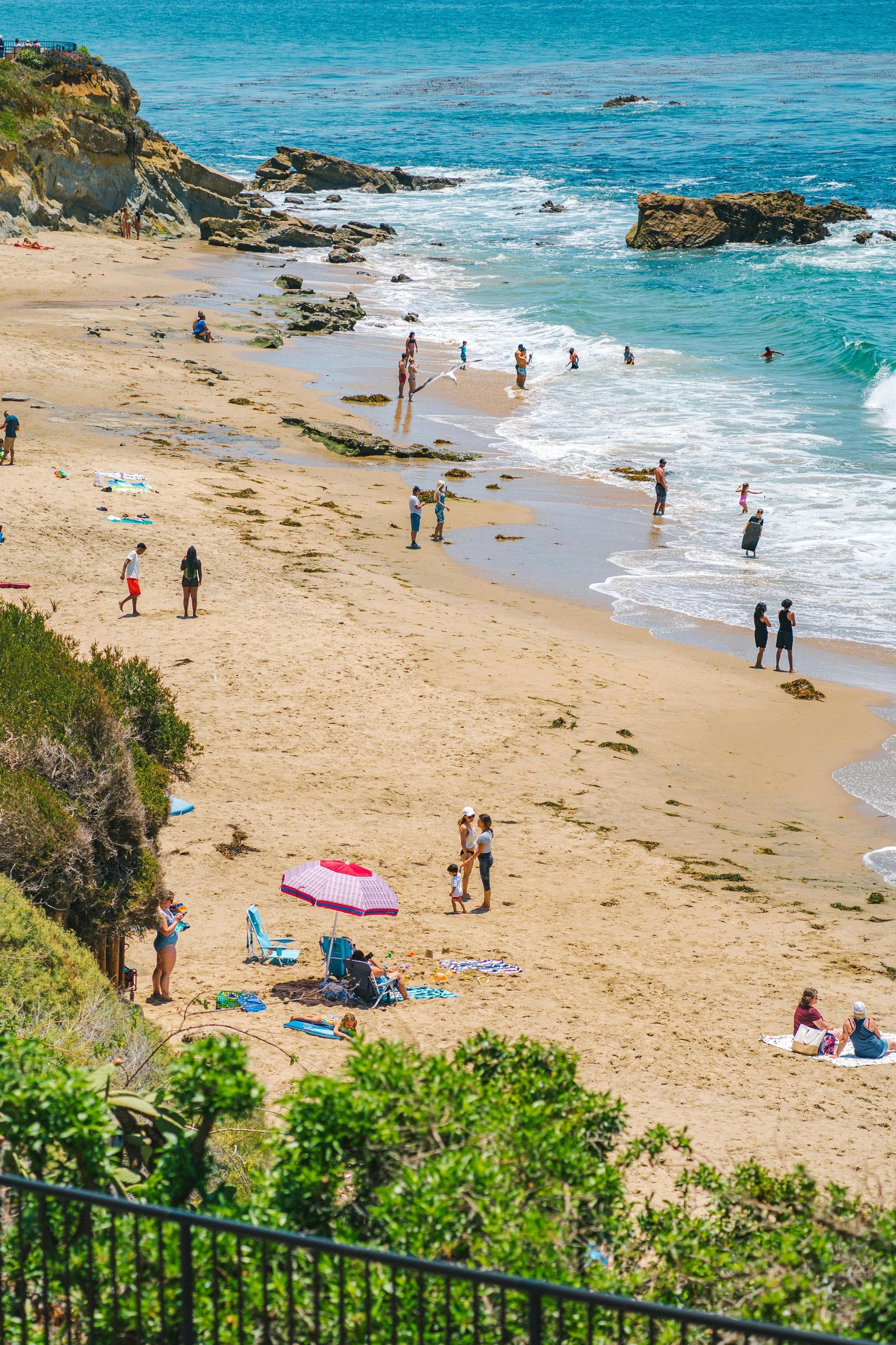 Rocky beach with people swimming, sunbathing, and walking near turquoise water on a sunny day