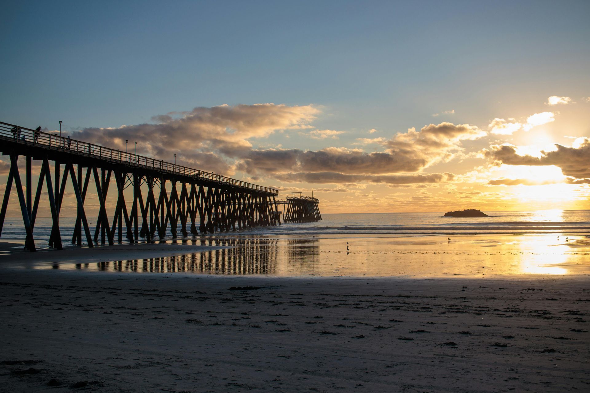 Sunset over a beach with a long pier and glowing reflections on the wet sand