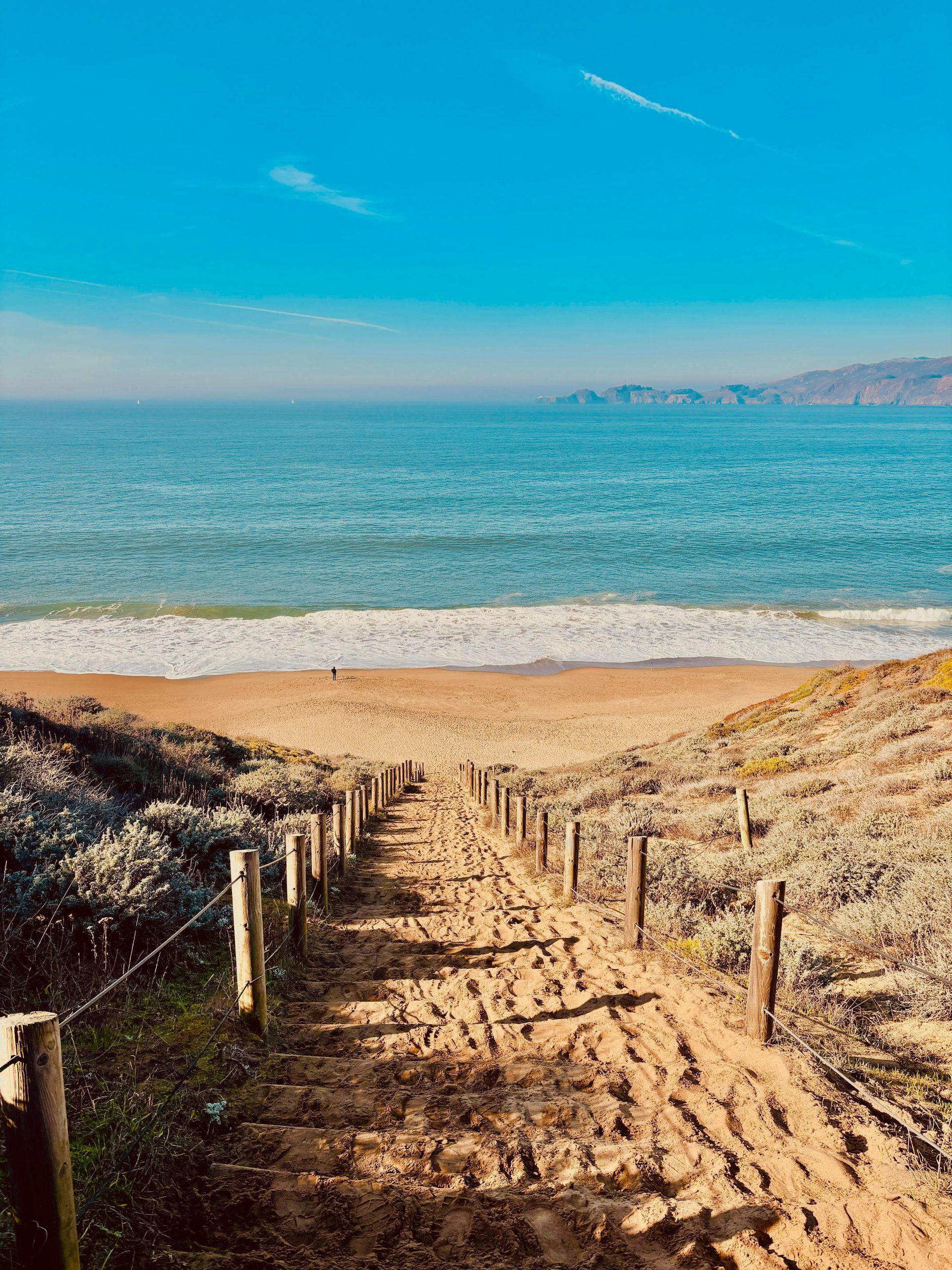 Sandy path leading to a calm blue ocean with a clear sky and distant mountains.