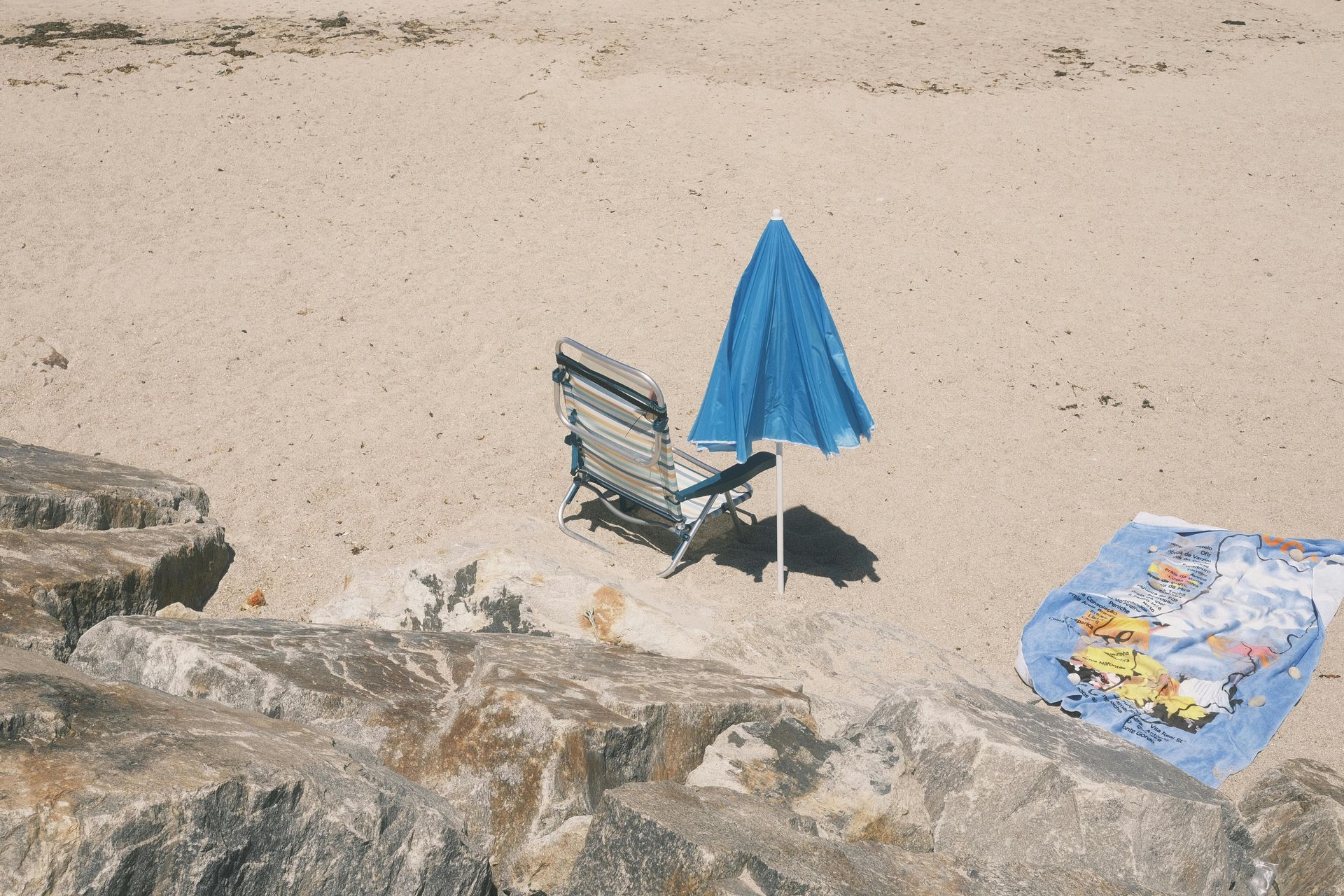 Blue beach umbrella and striped chair on a sandy beach beside a colorful towel.