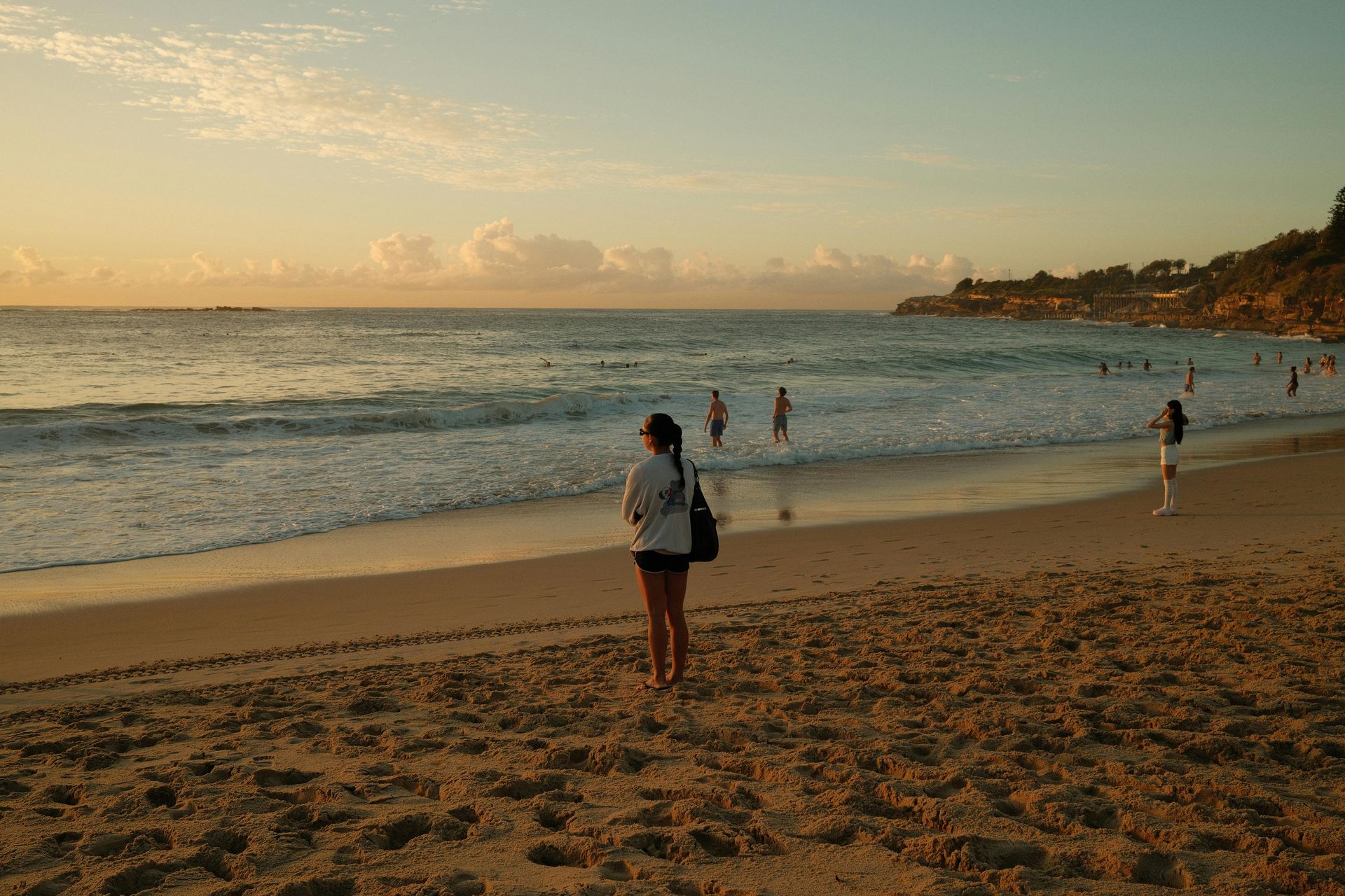 People standing on a sandy beach at sunset, with waves and a distant shoreline.