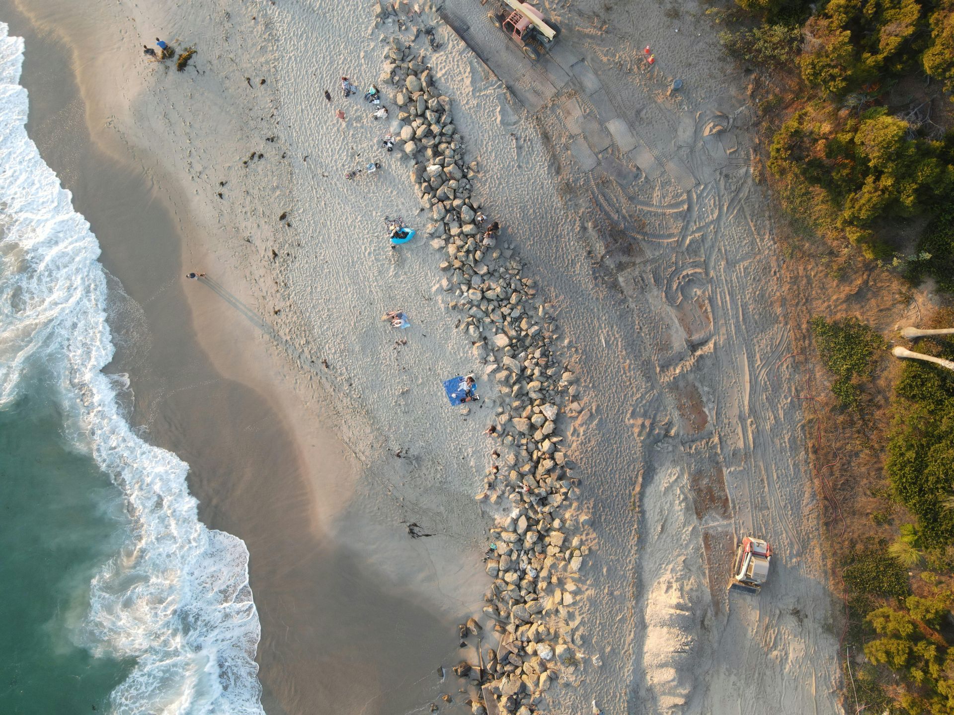 Aerial view of a rocky beach with waves, two blue umbrellas, and a dirt road along the shore