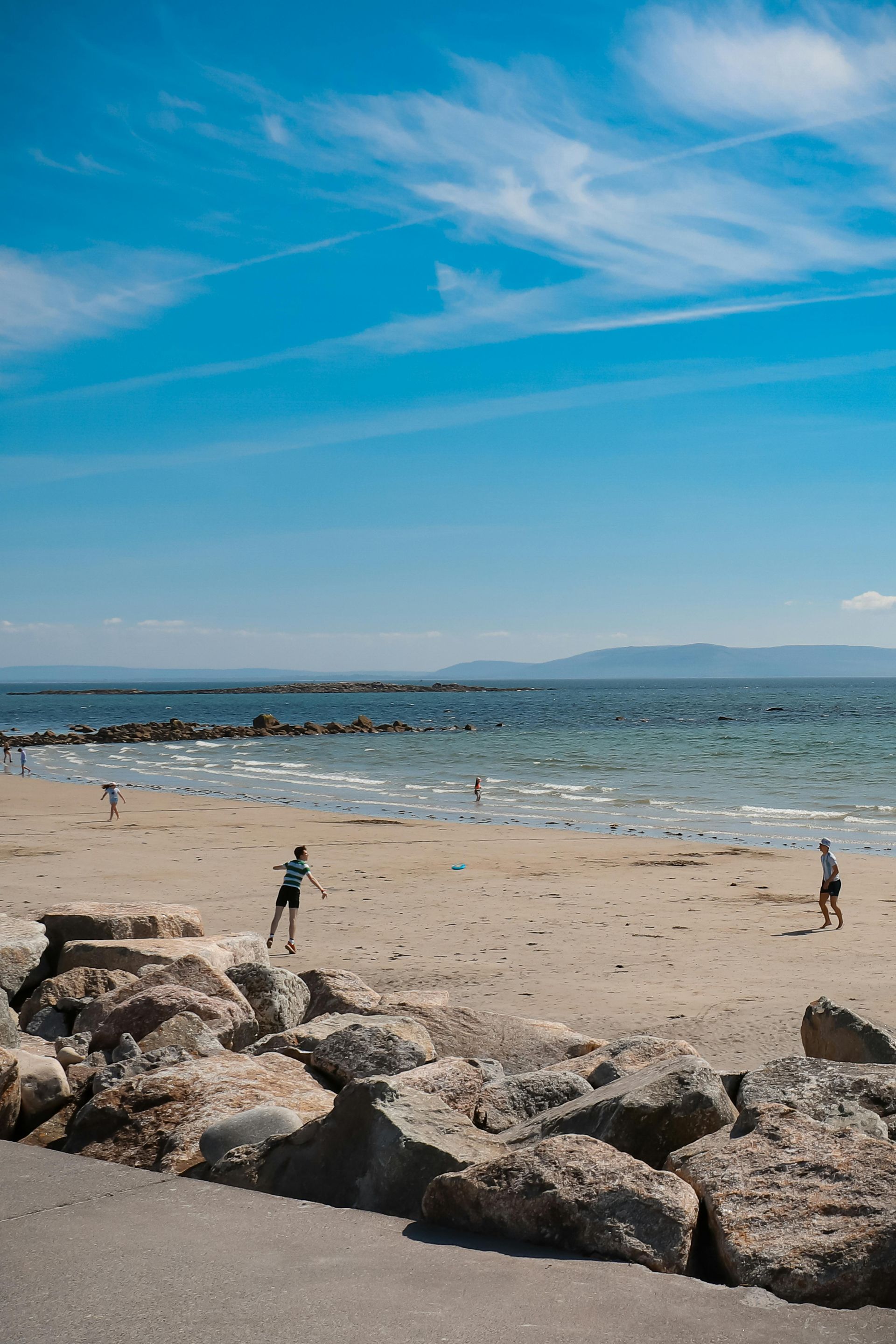 Rocky beach with two people near the shore, calm blue water, distant hills, and a bright sky with wispy clouds