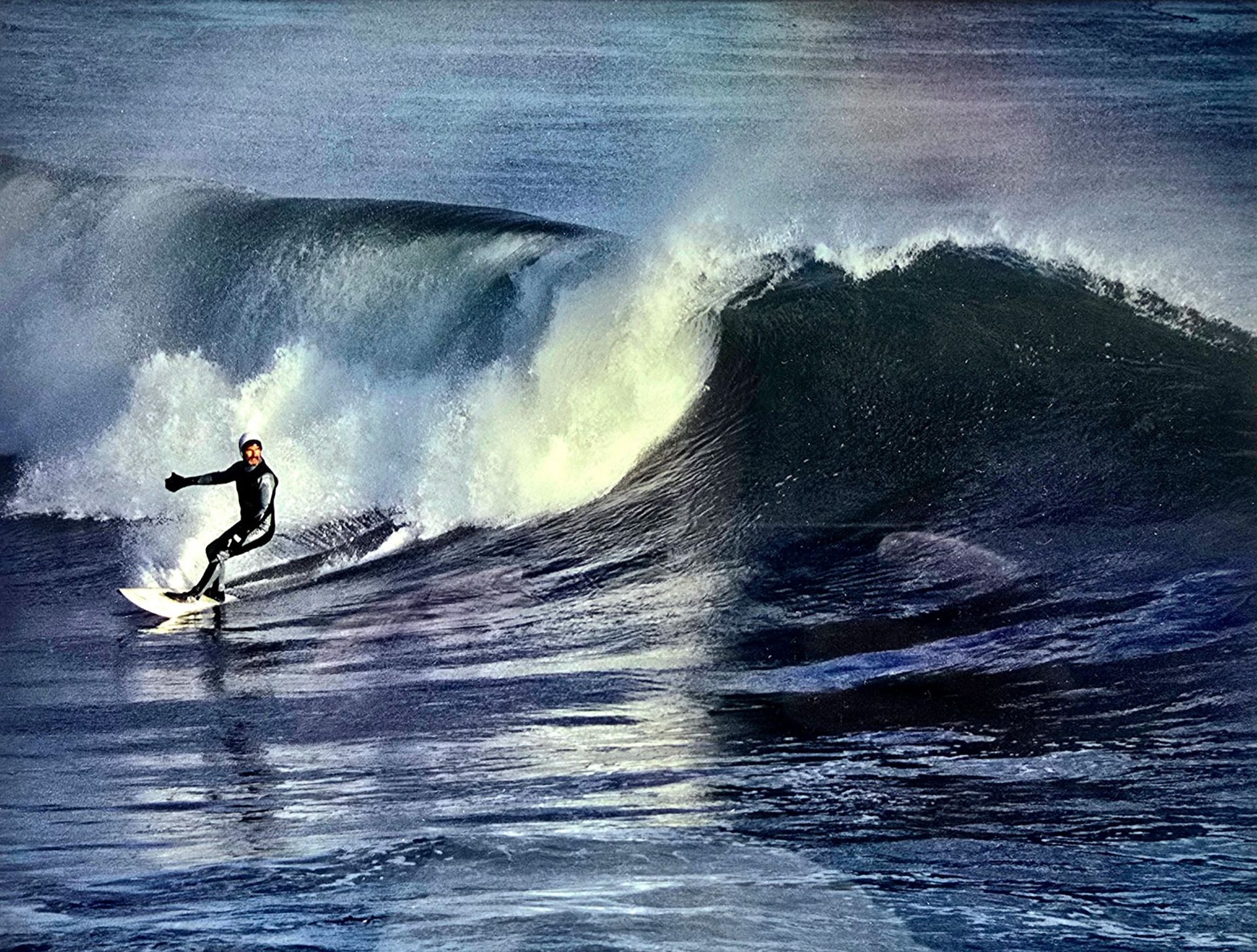 Surfer riding a large blue wave with spray breaking behind them